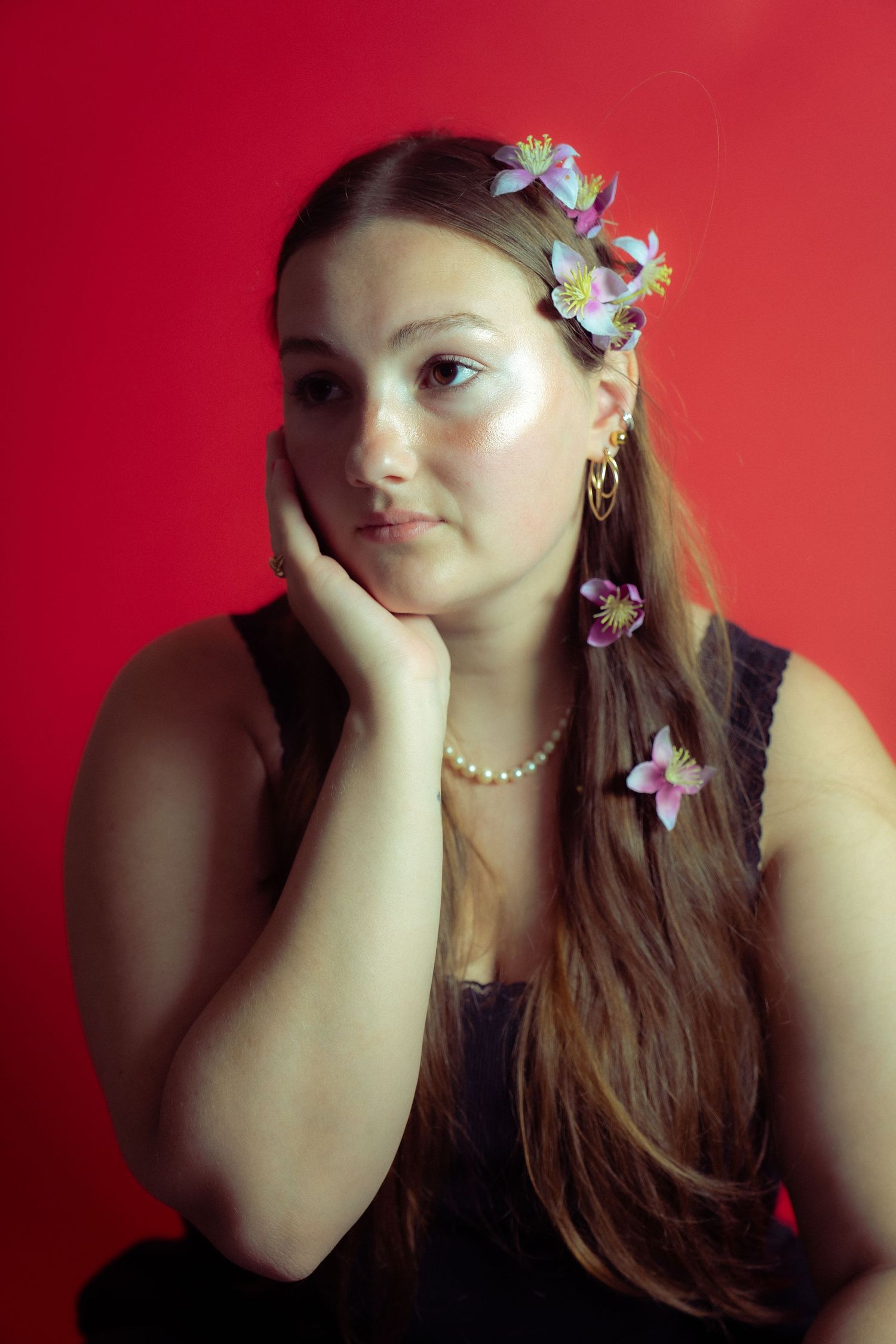 Portrait against red background featuring long brown hair adorned with delicate pink floral accessories and pearl necklace.