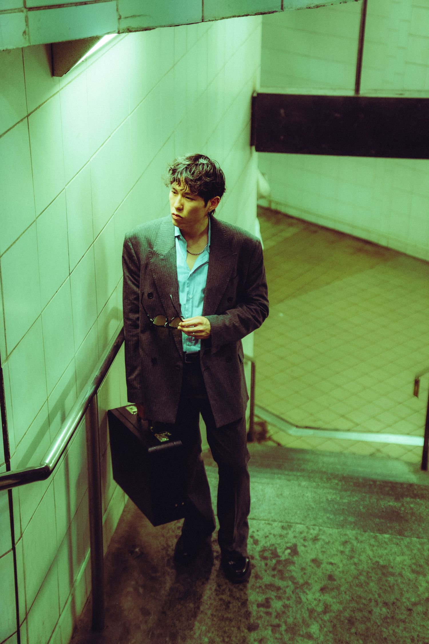 Young man in dark suit holding briefcase stands in green-lit subway stairwell, looking contemplative.