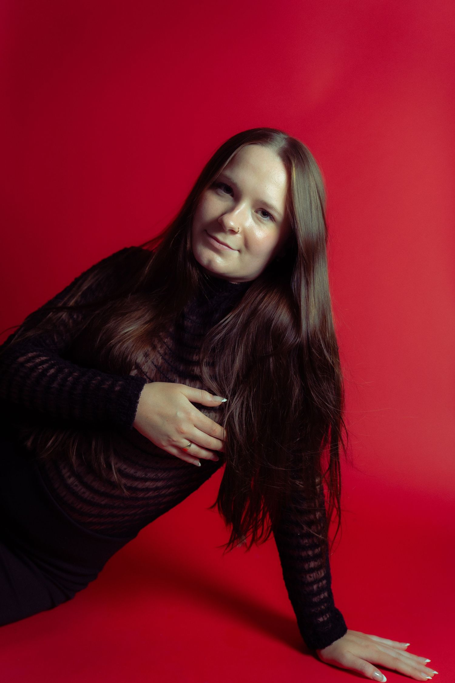 A person with long dark hair poses playfully against a vibrant red background while wearing a black striped top.