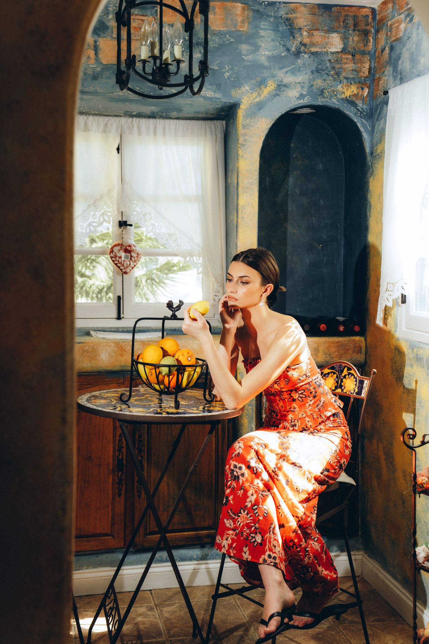 Woman in red floral dress sitting at ornate table with lemons in vintage Mediterranean-style interior.