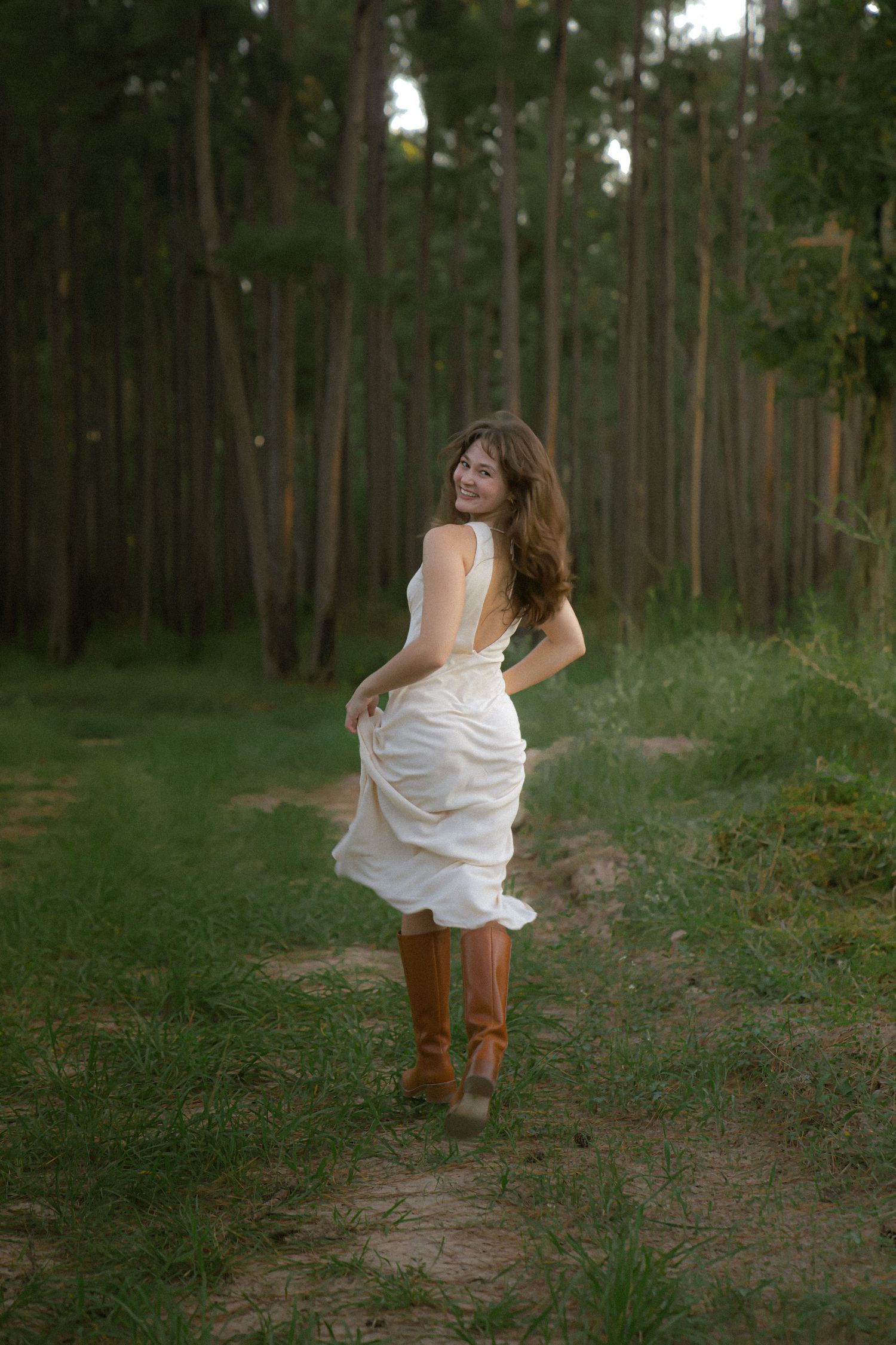 Woman in white dress and brown boots walking along a forest path at sunset.