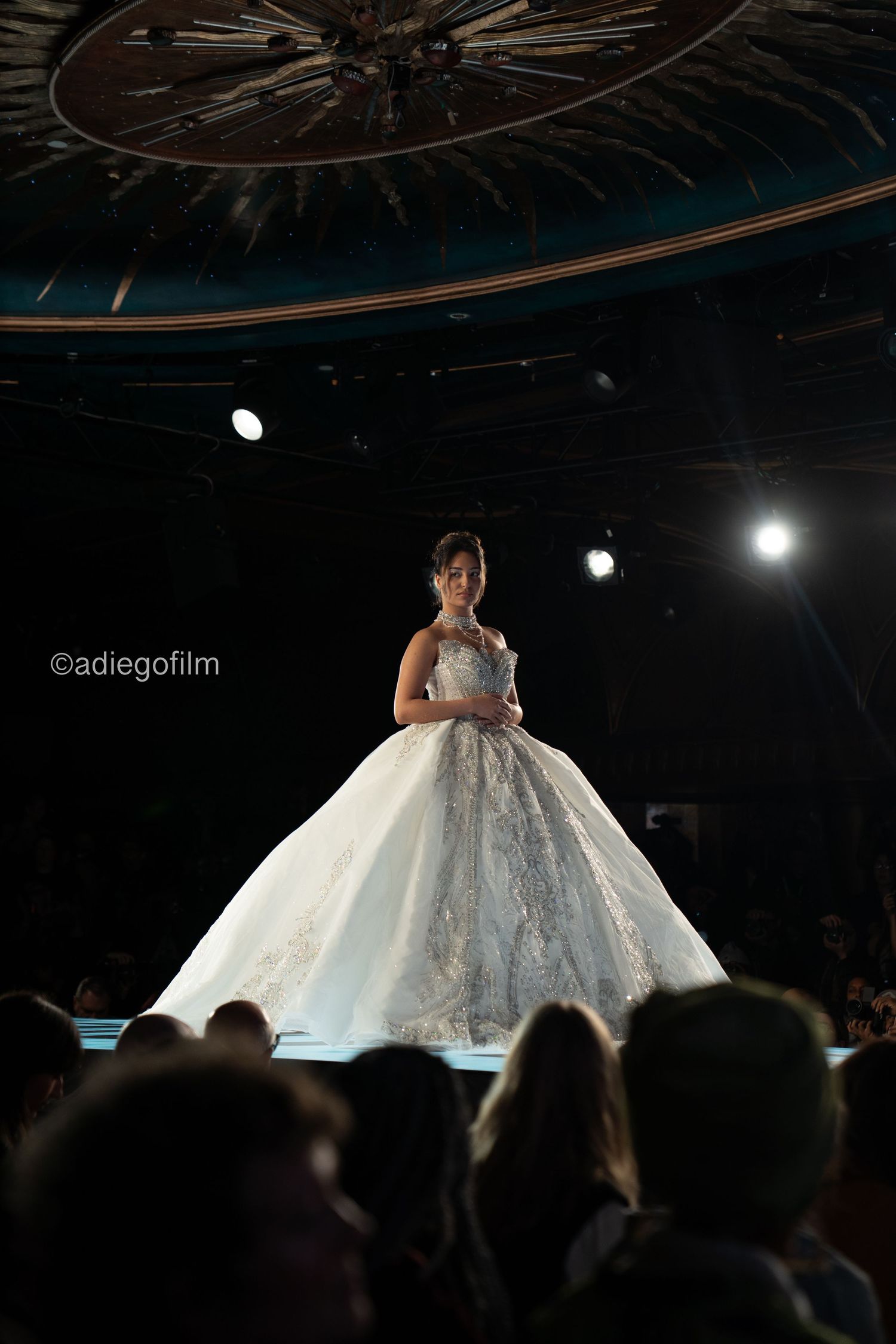 A model showcases a stunning white ball gown wedding dress on a dark runway with dramatic spotlighting.