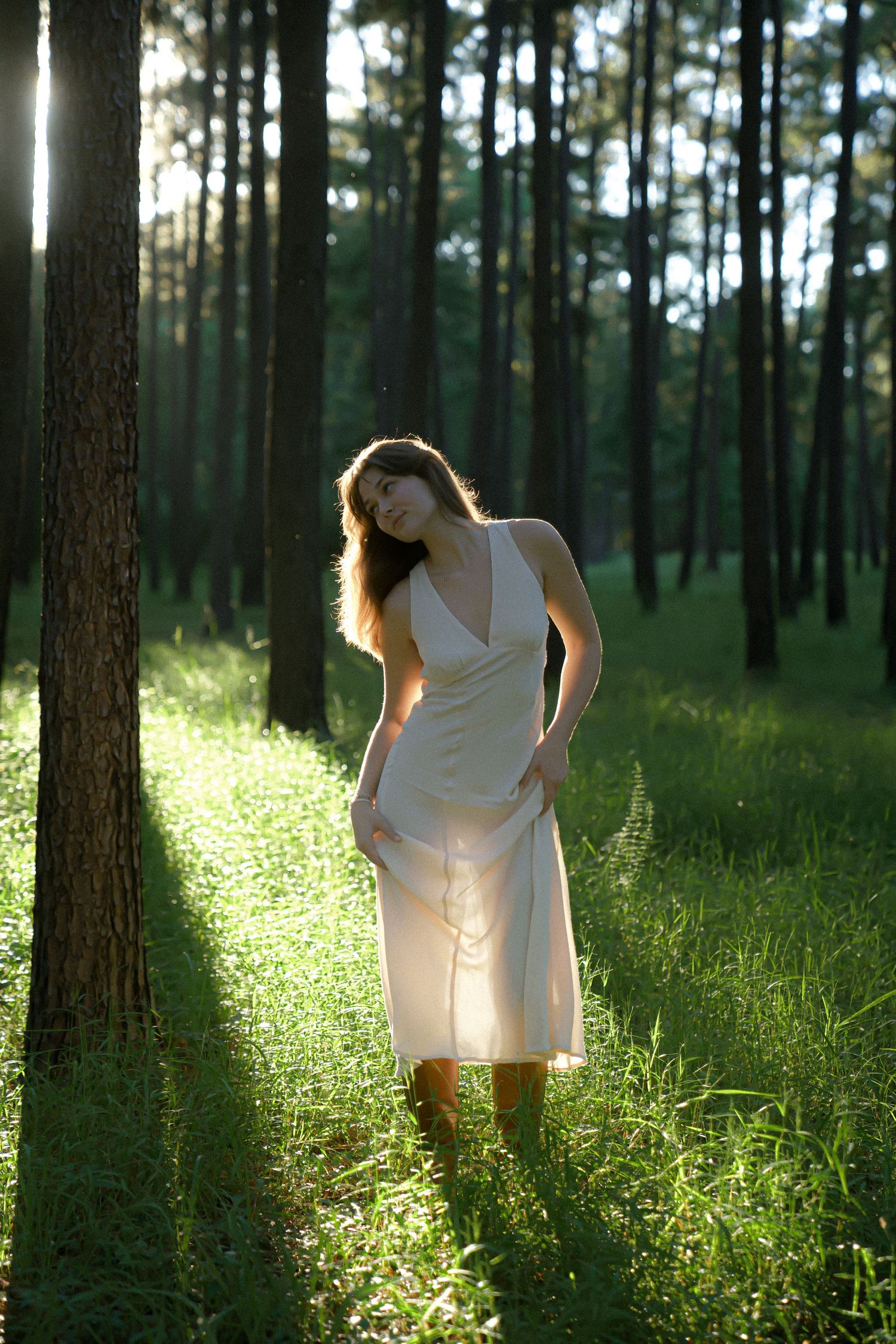 White dress flowing in dappled sunlight between tall pine trees in a tranquil forest.