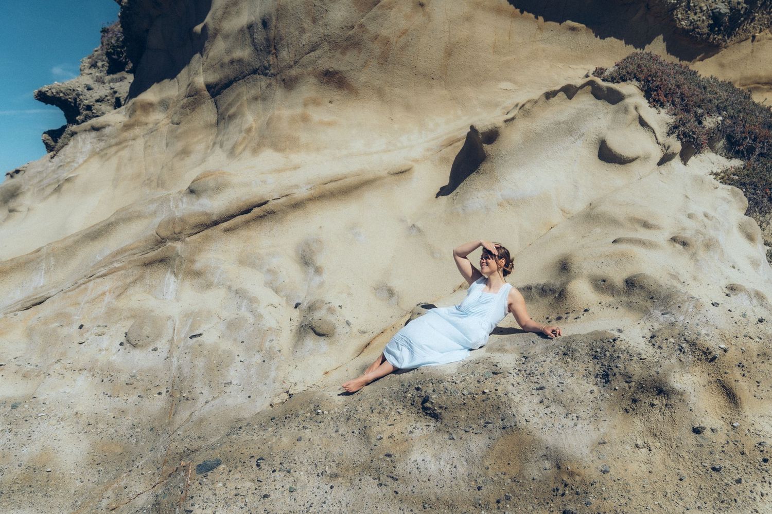 Woman in white dress reclining on sandy rocky cliffs under a bright blue sky.