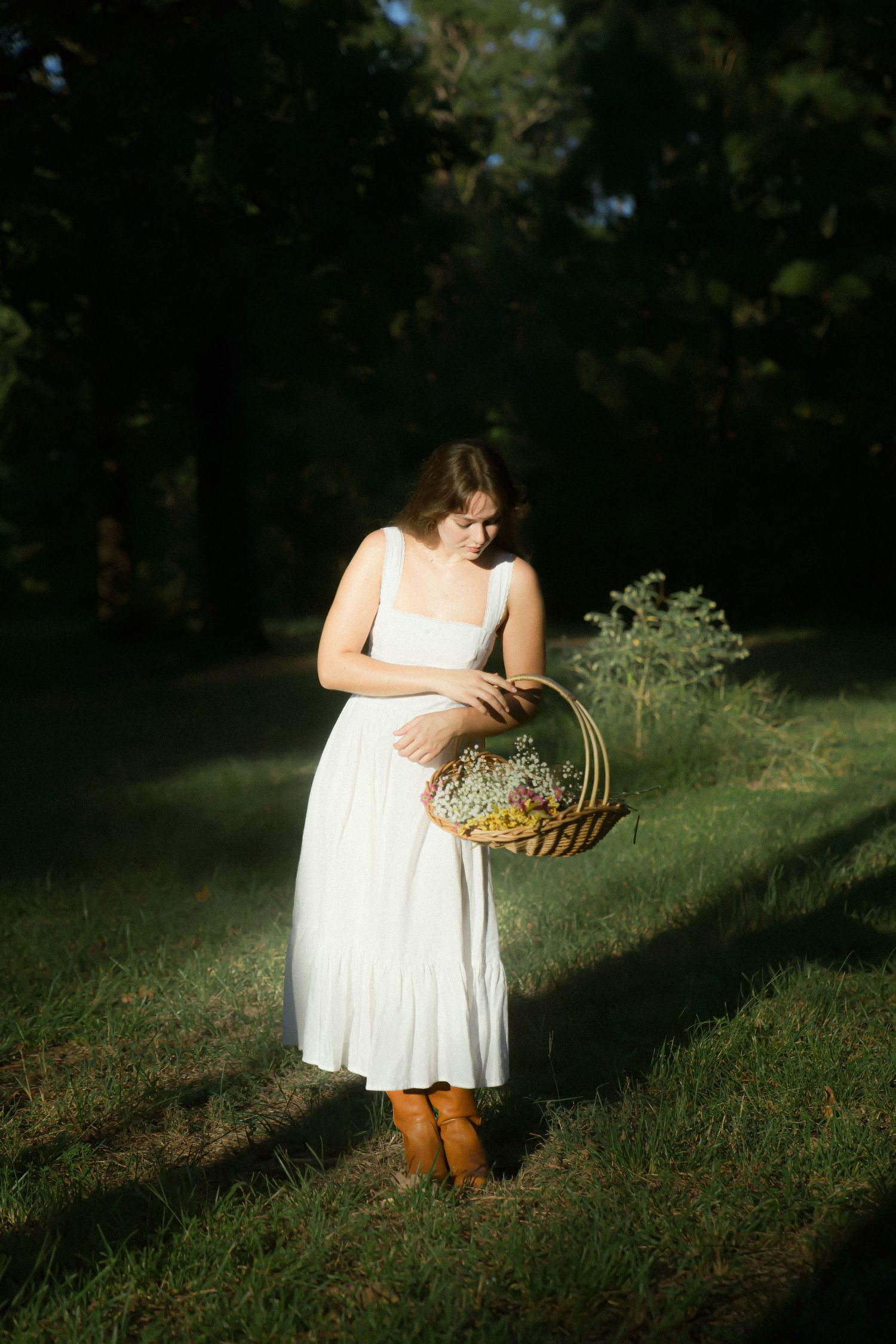 Figure in white summer dress holding woven basket stands in dappled sunlight on grassy forest path.