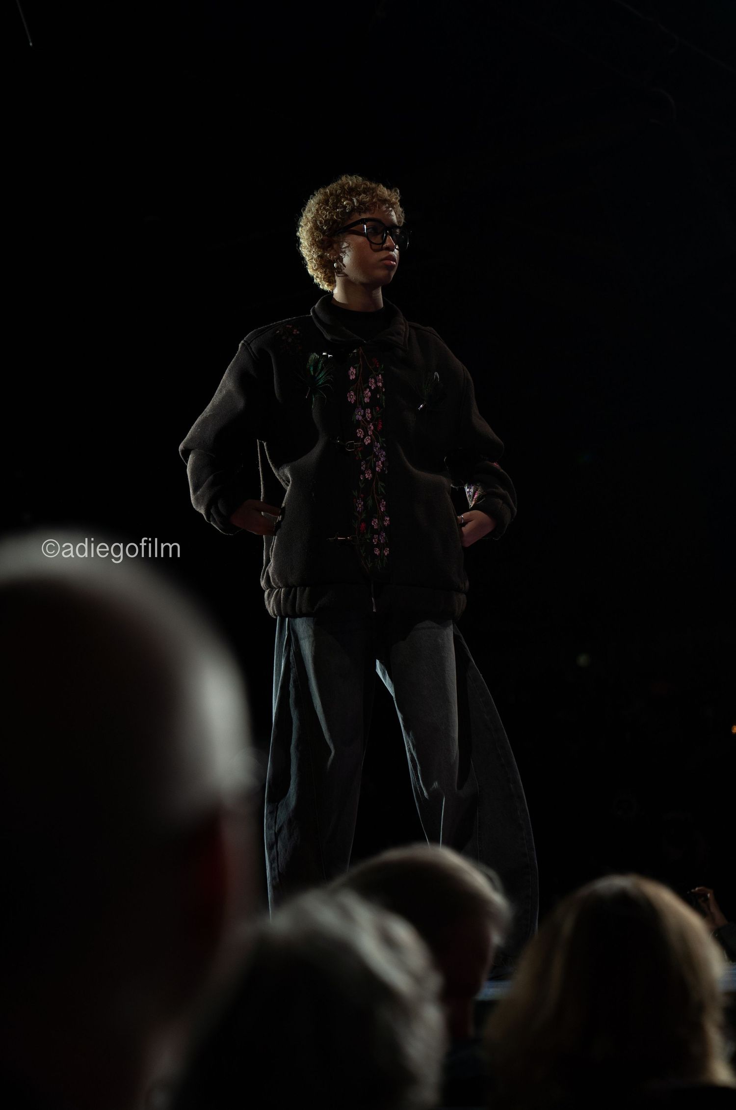 Figure in black clothing stands on stage performing in dramatic lighting against dark background.