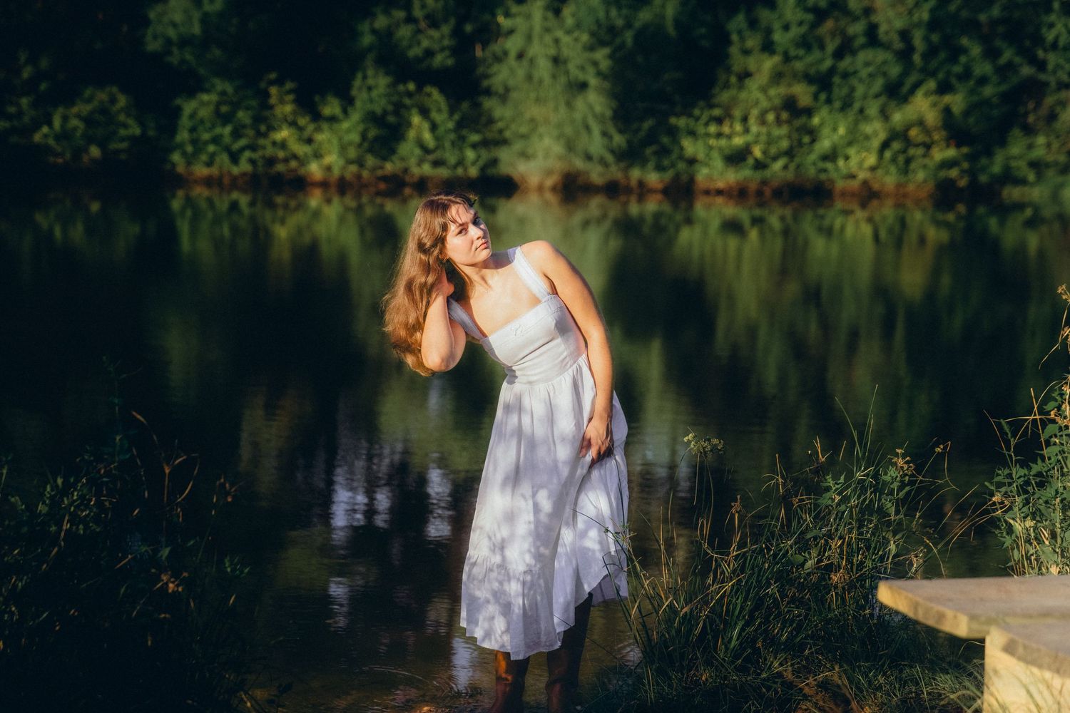 Graceful pose in white dress against serene lake and forest backdrop during golden hour.