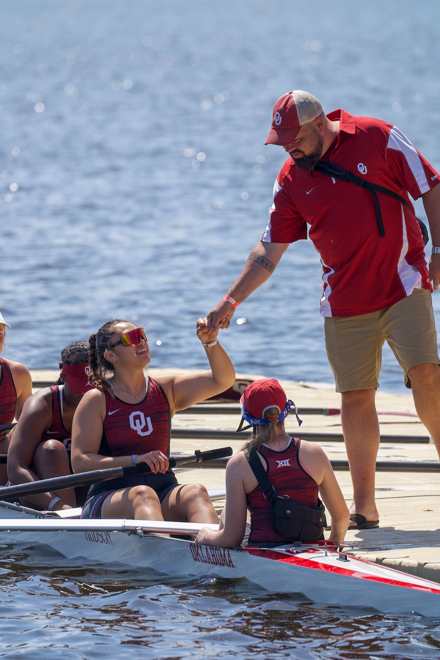 A rowing coach in a red jacket fist bumps an athlete in a boat on sparkling water by the dock.