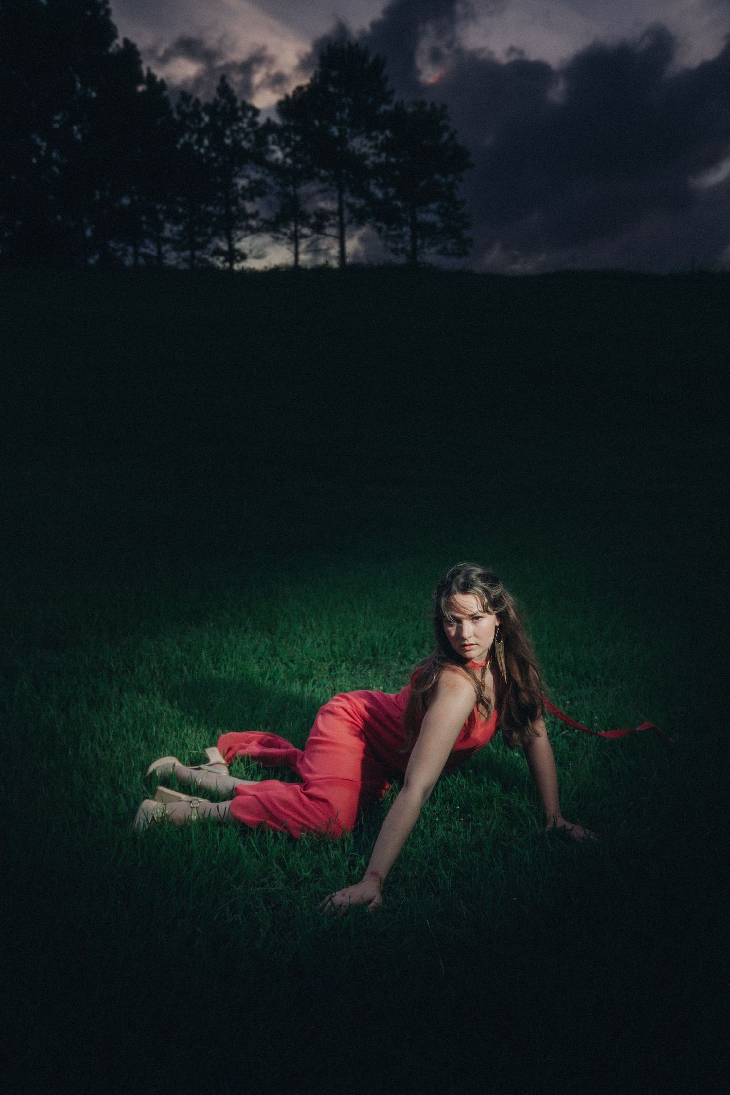 Woman in red dress reclining on lush green grass at dusk with dramatic cloudy sky and tree silhouettes.