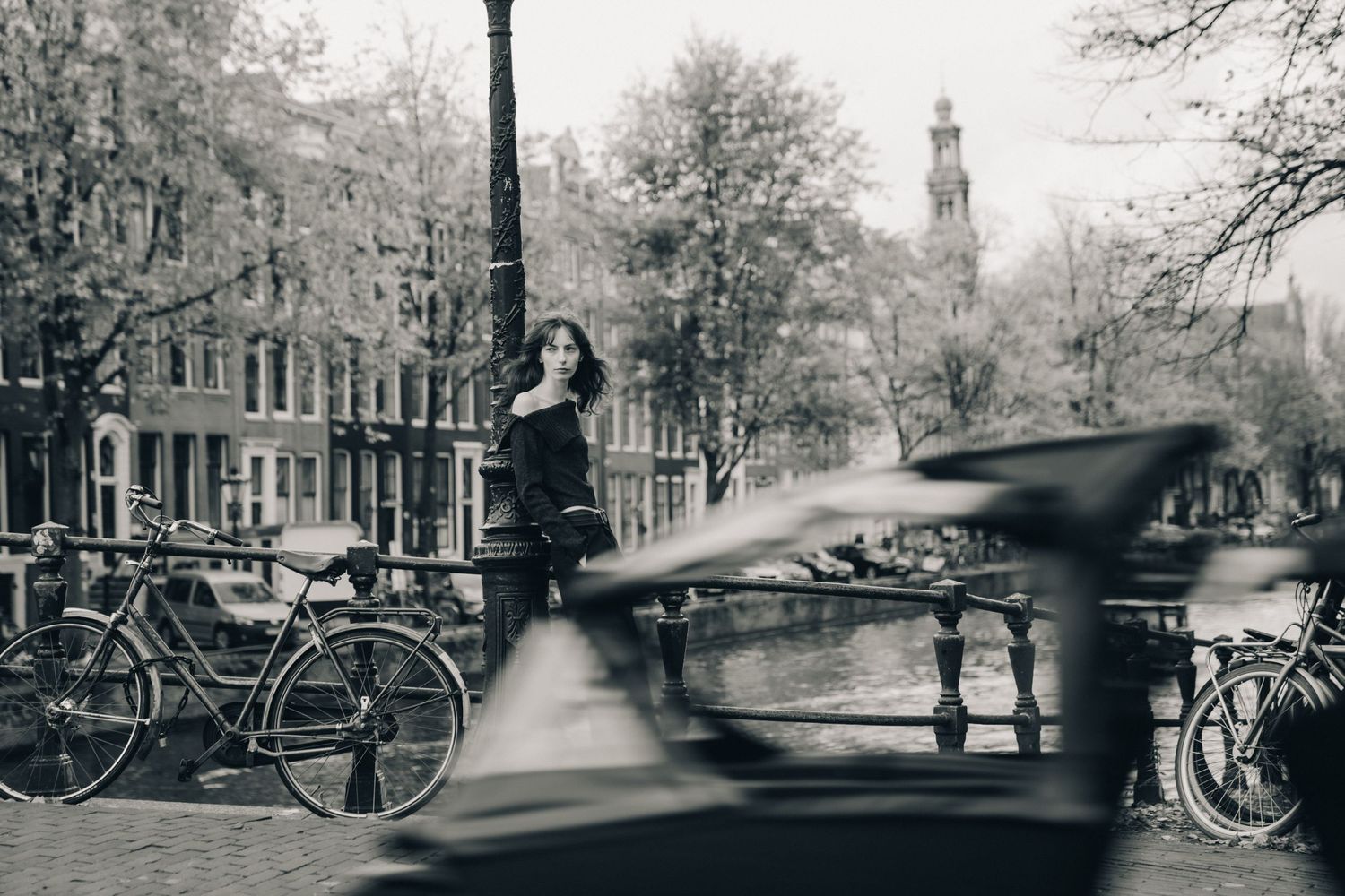 A black and white photograph of a canal bridge in Amsterdam with bicycles parked along the railing and trees in background.