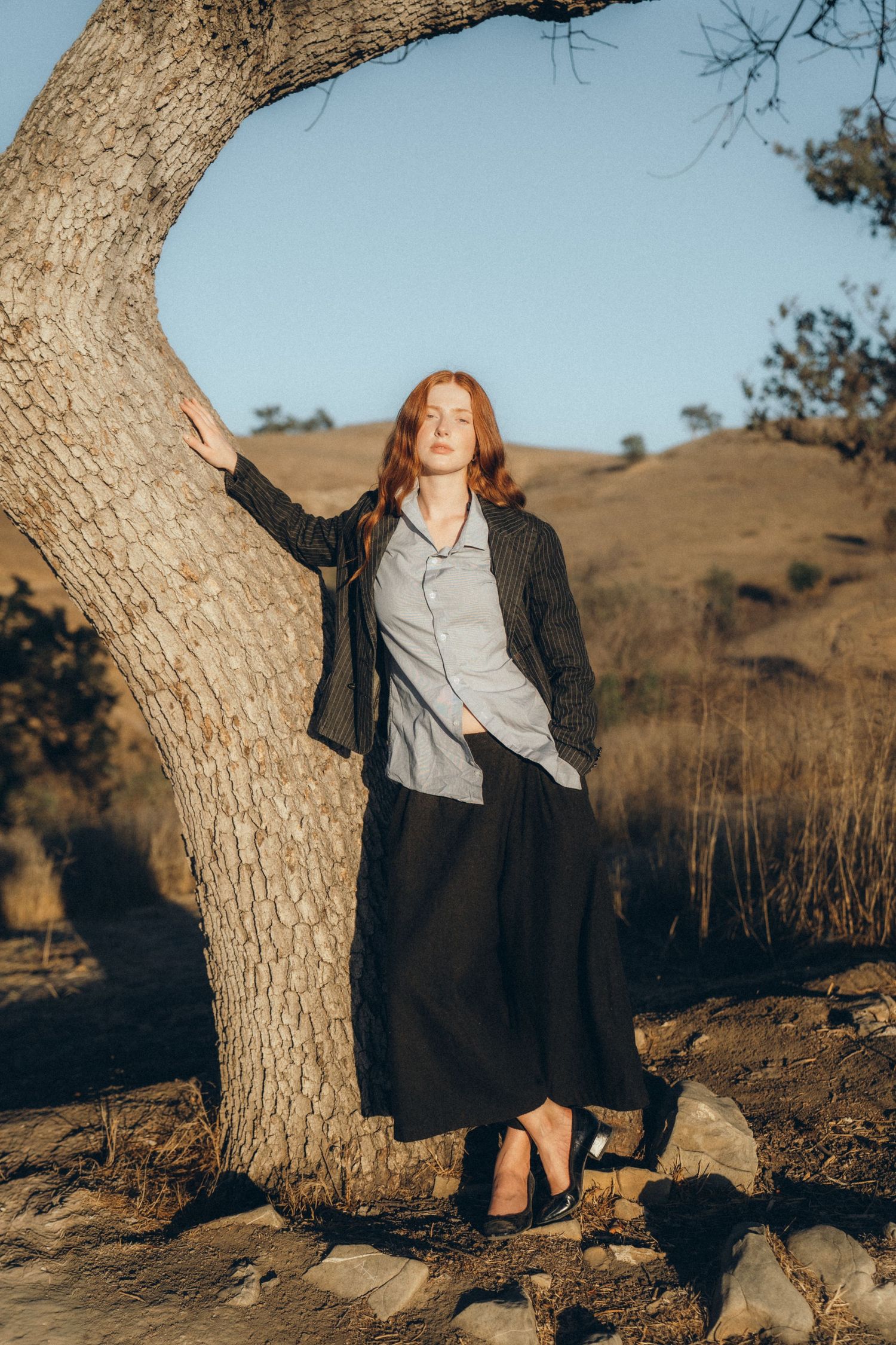 A person in a long black skirt and gray blouse leans against a tree trunk in a rural hillside setting at golden hour.