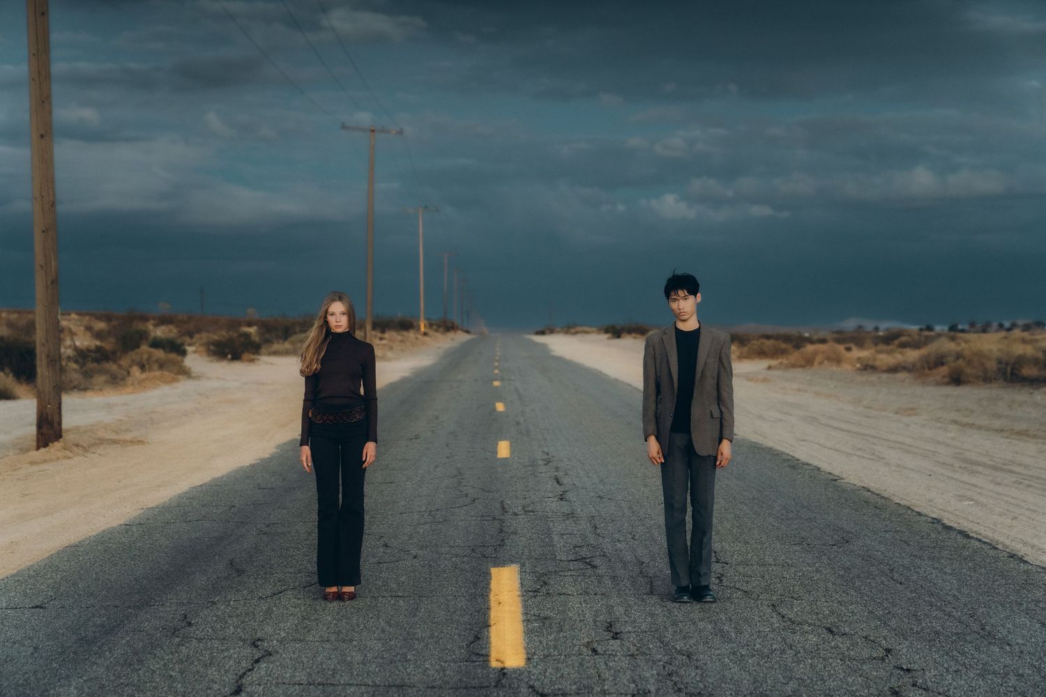 A person stands alone on a long empty desert road stretching into the horizon under a dramatic stormy sky.