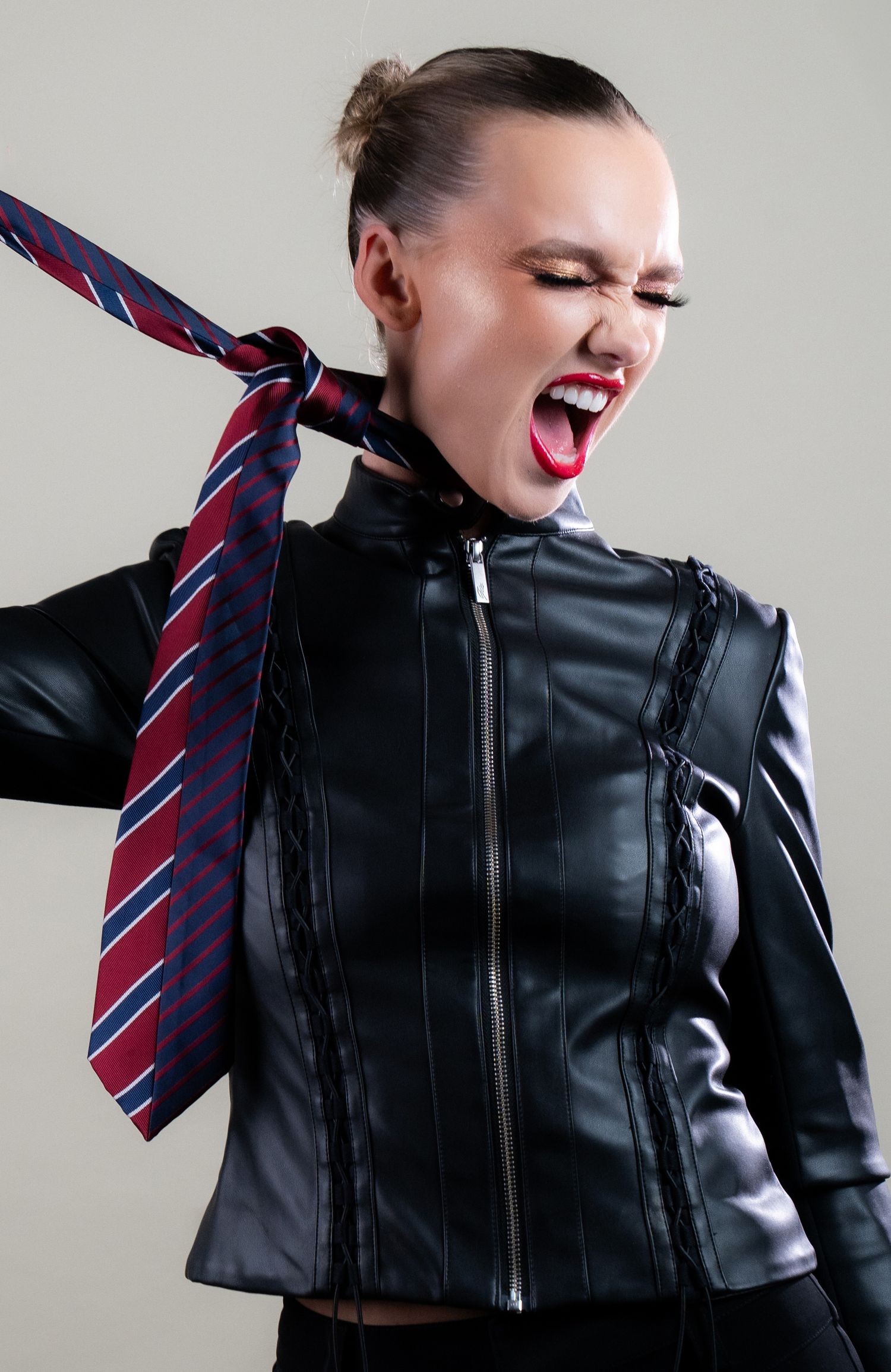 Woman in black leather jacket with red lips, pulling a striped tie around her neck, mouth open in a scream.