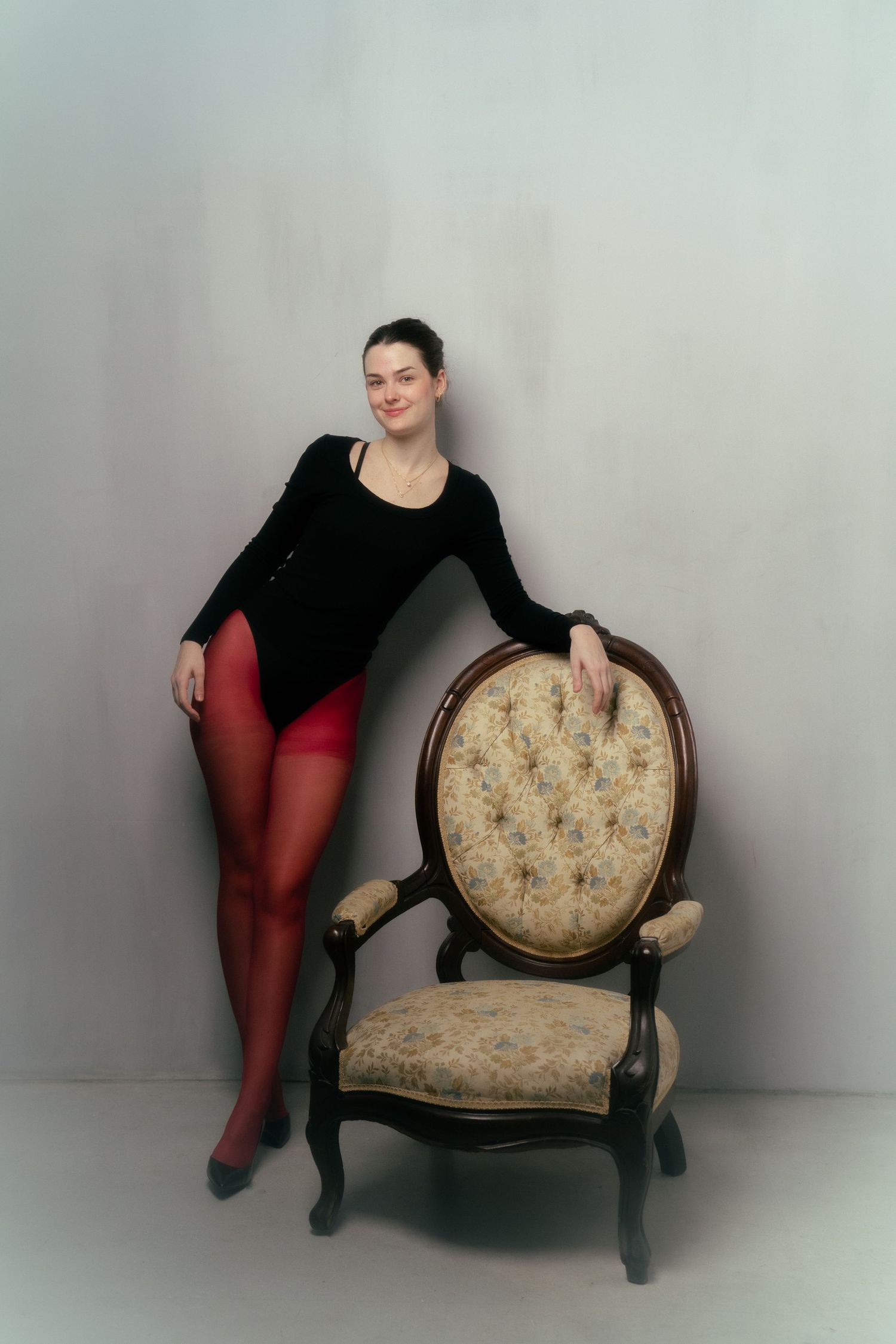 Woman in black top and red tights posing beside an ornate vintage armchair against a gray background.