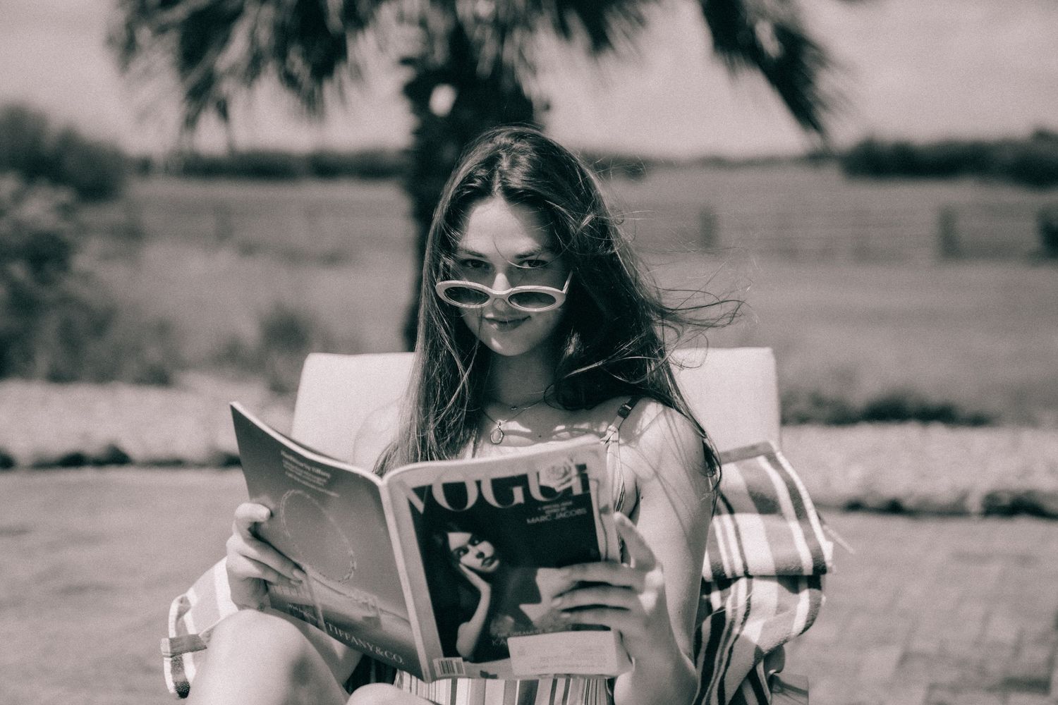 Someone wearing sunglasses reads a vintage magazine outdoors in a black and white photograph.