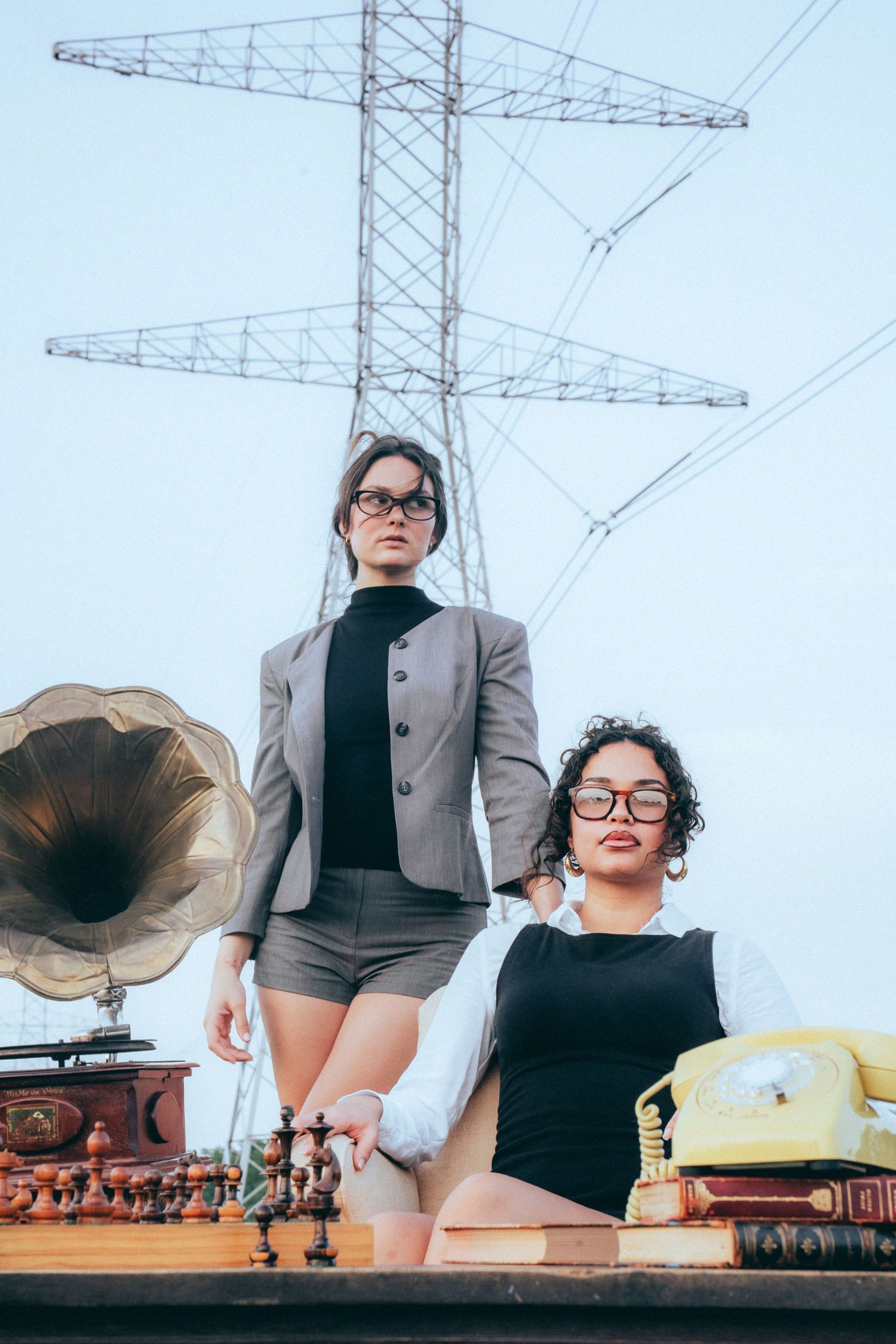 Two models pose on vintage furniture with retro items including phonograph and telephone against industrial power line tower.