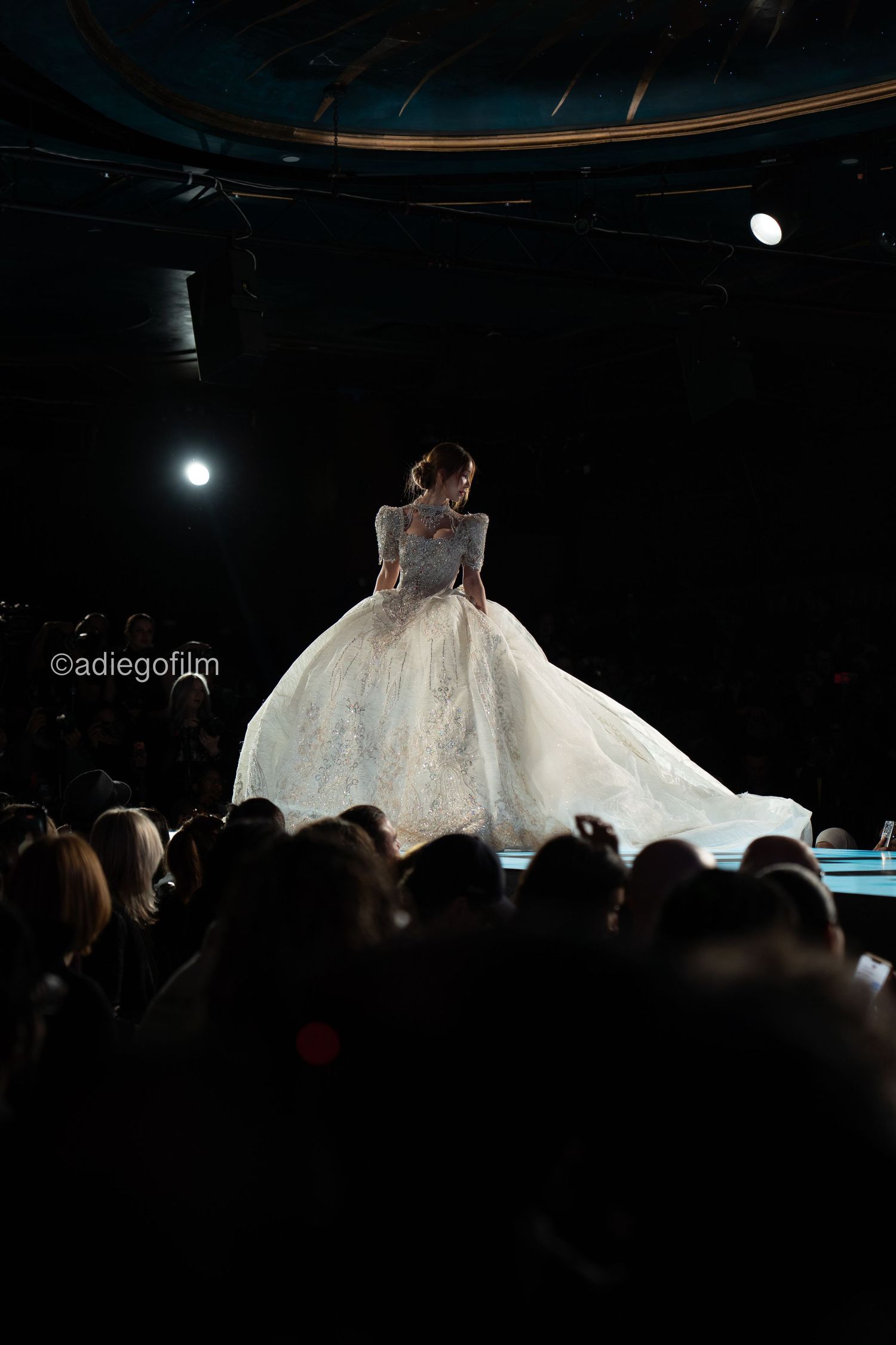 A dramatic back view of a voluminous white wedding gown being presented on a dimly lit fashion runway stage.