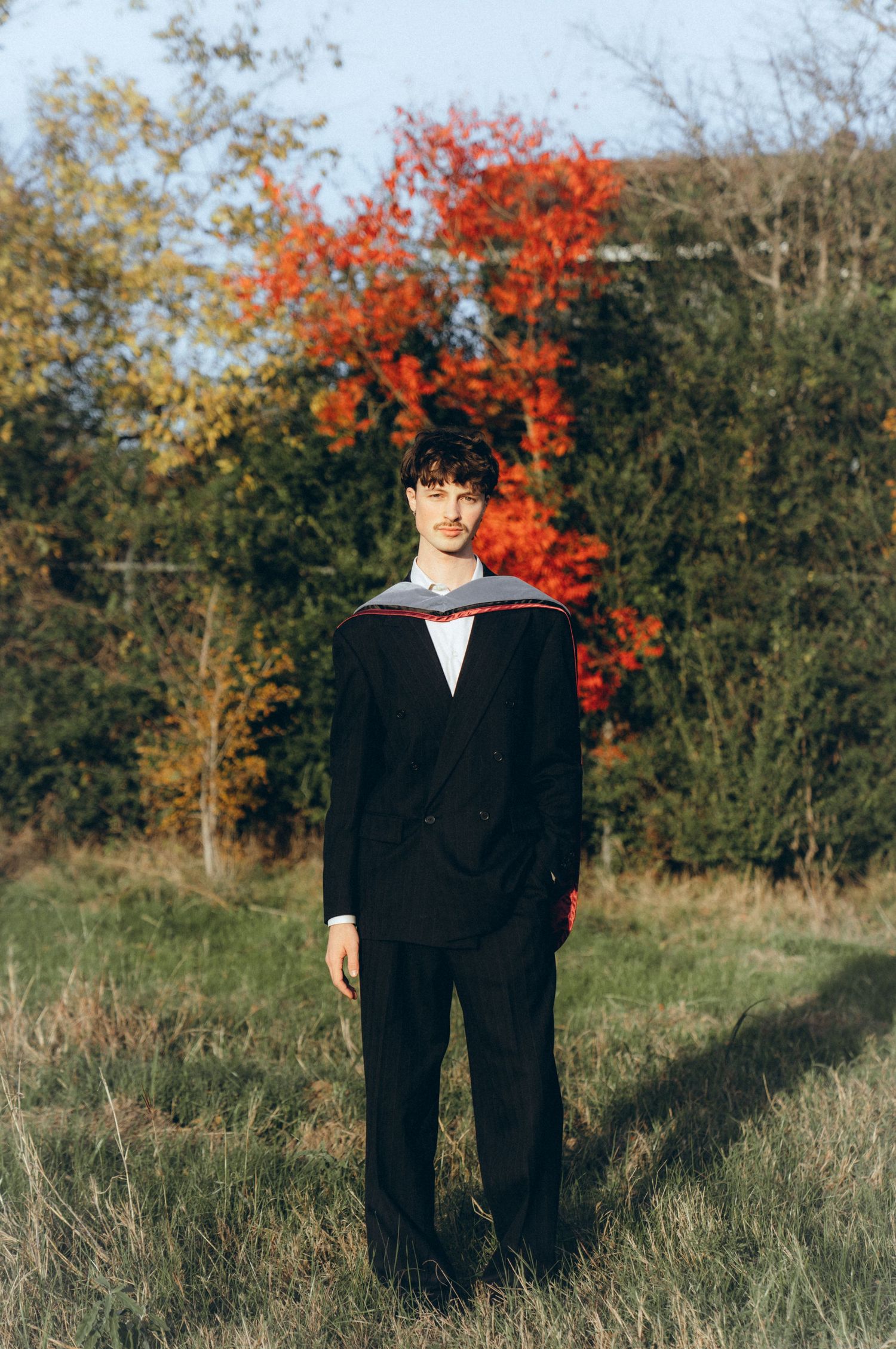 Two people in black coats stand together against a backdrop of vibrant autumn foliage and trees.