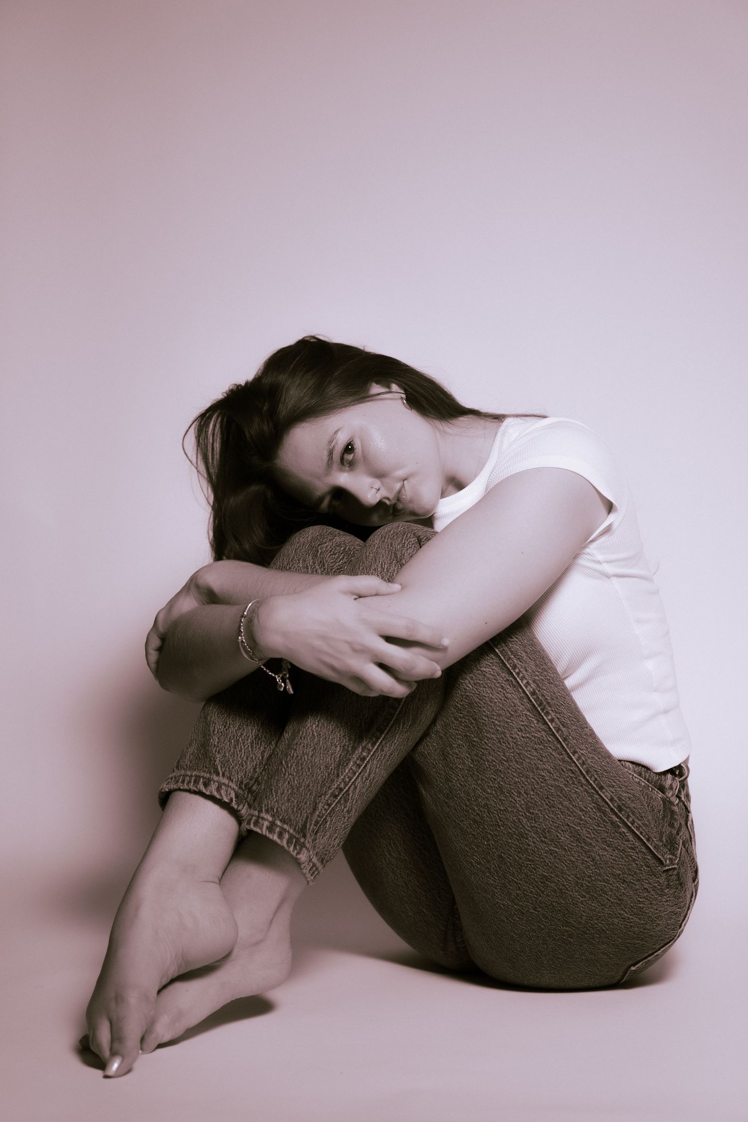 A young person in white shirt and jeans sits curled up against a neutral colored wall with a contemplative expression.