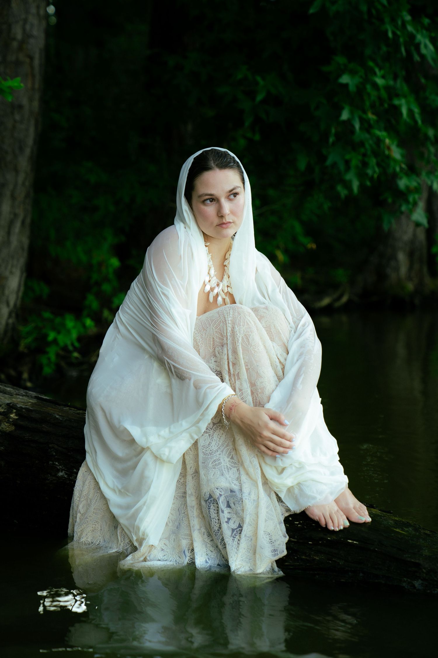 Figure in ethereal white dress and veil sits contemplatively by a dark pond in a lush forest setting.