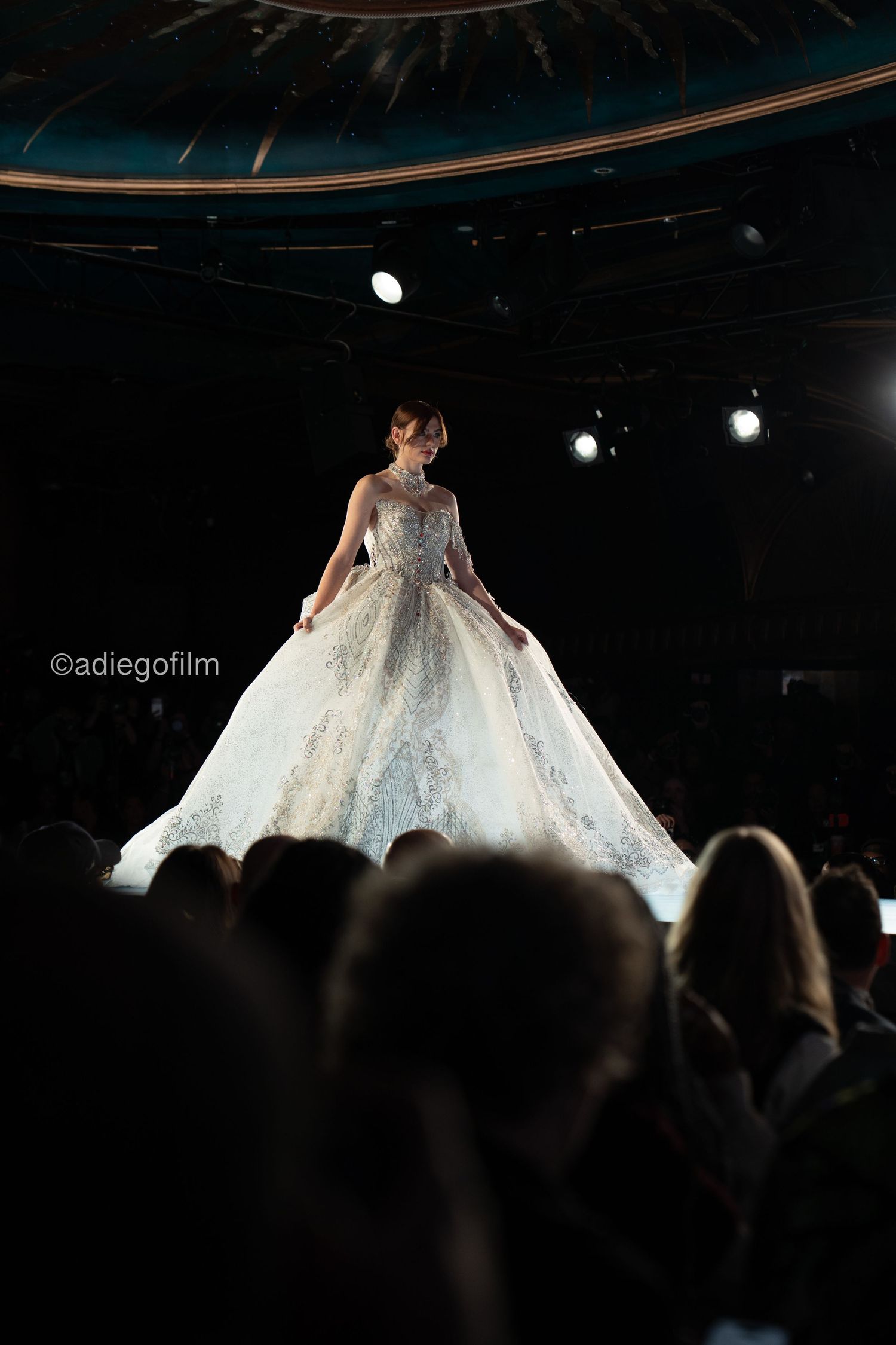 A model walks down a dark runway showcasing an elegant white wedding gown with a dramatic full skirt at a fashion show.