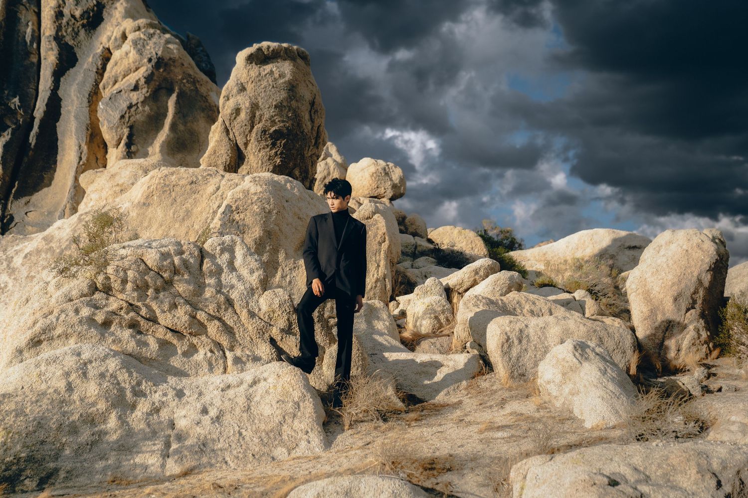 A silhouetted figure stands on rocky terrain against a dramatic cloudy sky at sunset.