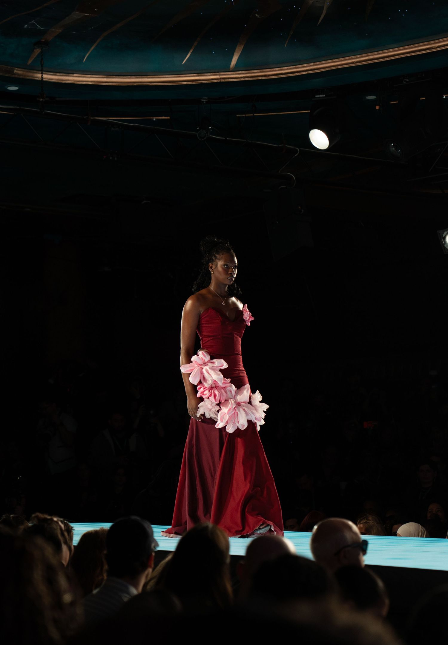 A performer in a burgundy dress walks gracefully across a dimly lit stage with pink flowers in hand.