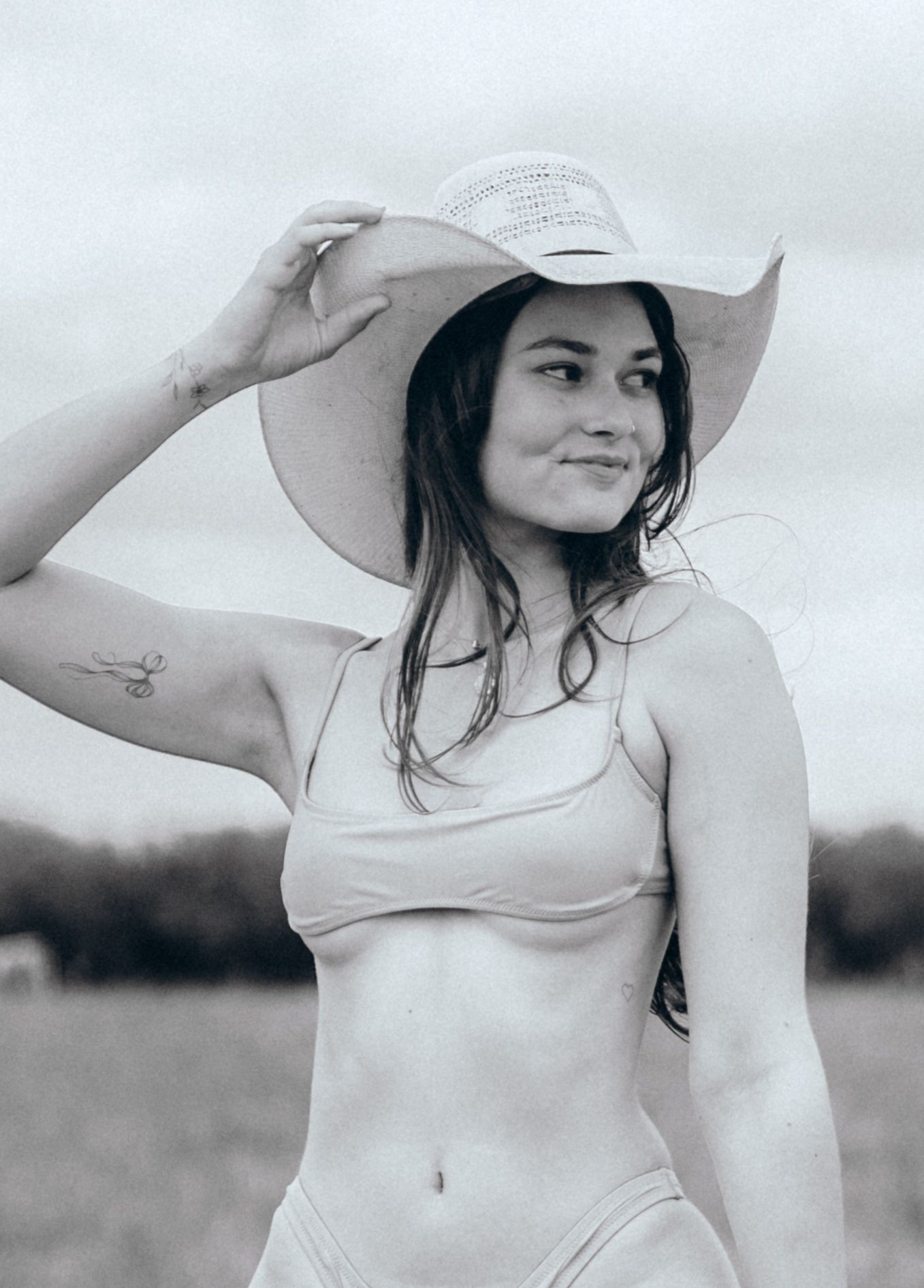 Black and white portrait of someone in a light sundress and wide-brimmed hat posing in an outdoor field.