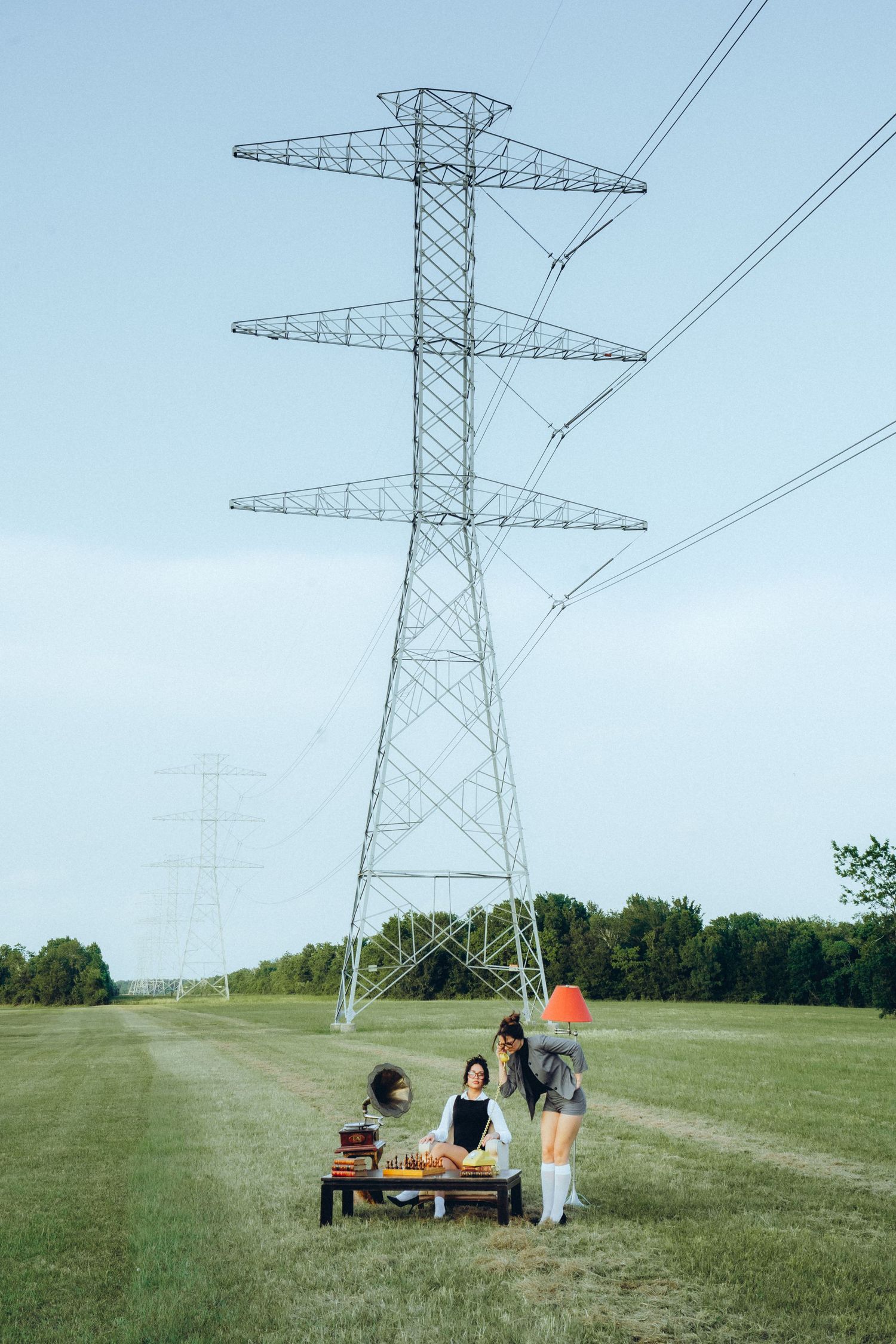 People gather at a picnic table beneath a towering electrical transmission tower in a grassy field on a sunny day.
