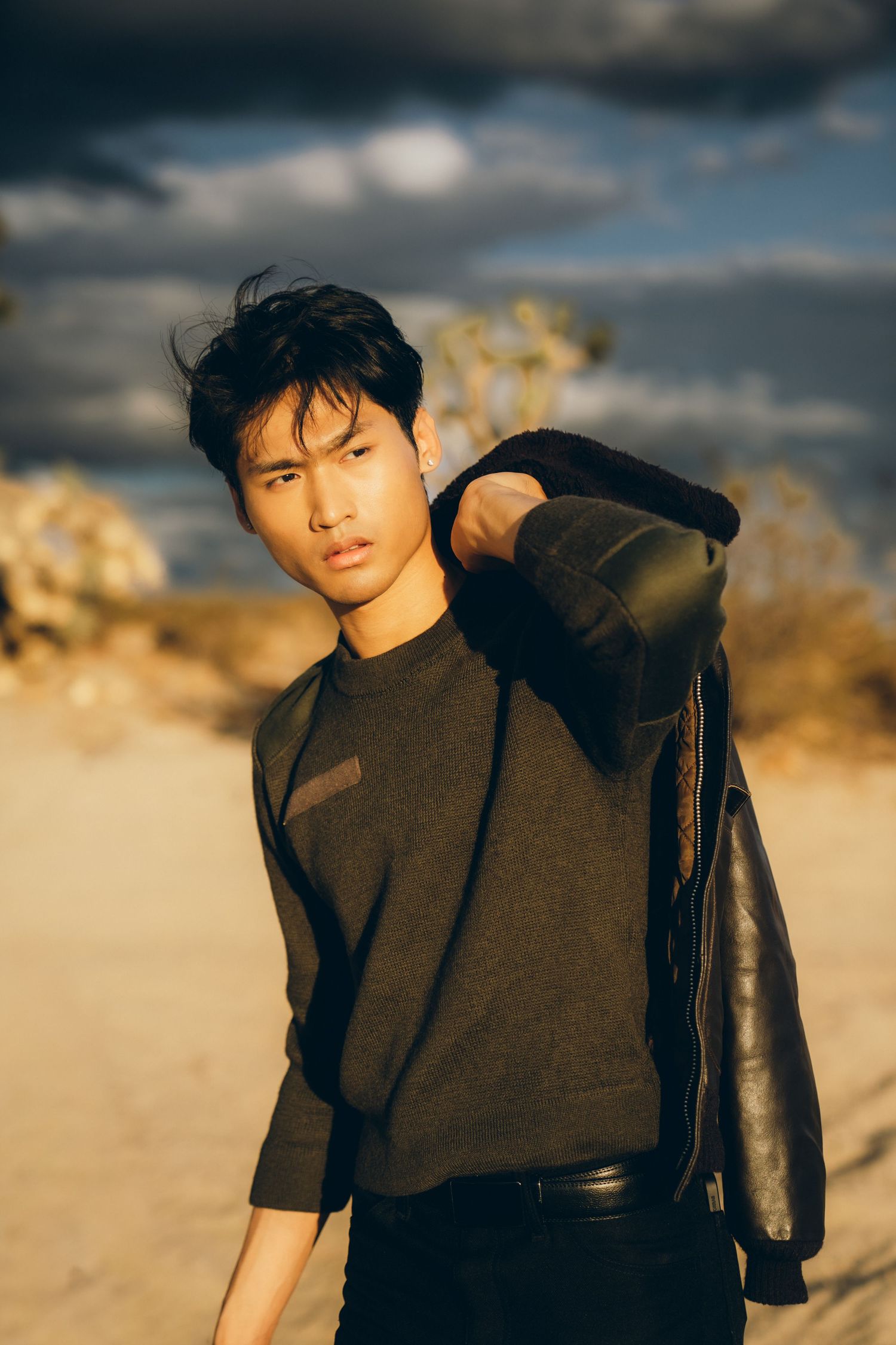 Fashion model wearing black leather jacket and dark clothing poses against desert landscape at golden hour.