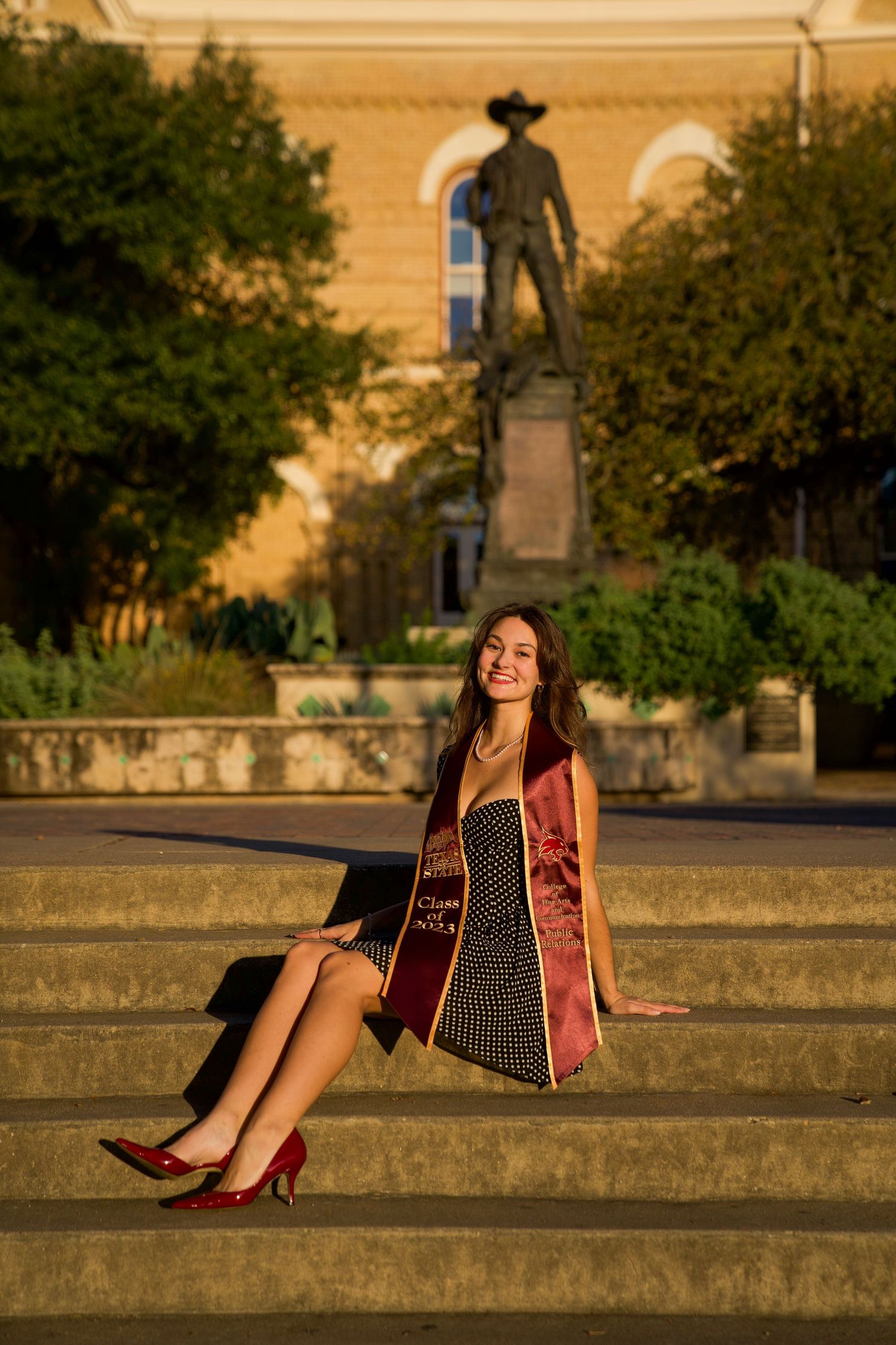 Graduate in red stole and polka dot dress sits on stone steps during golden hour.