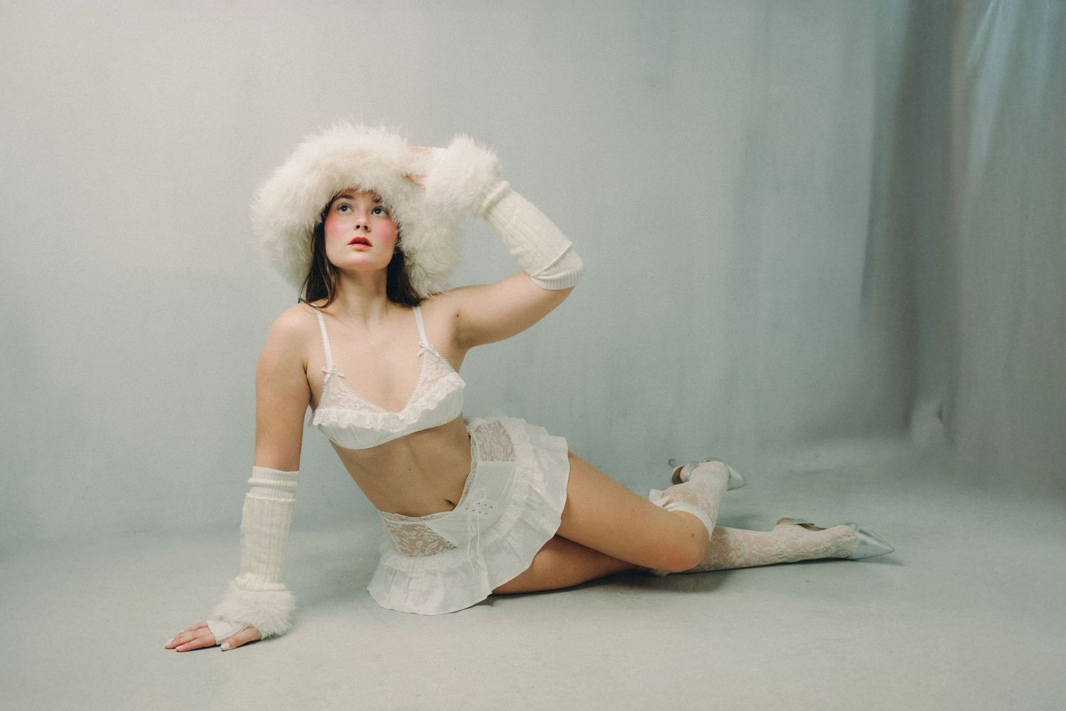 A person in white lingerie and long gloves poses gracefully on the floor against a soft, misty gray backdrop.