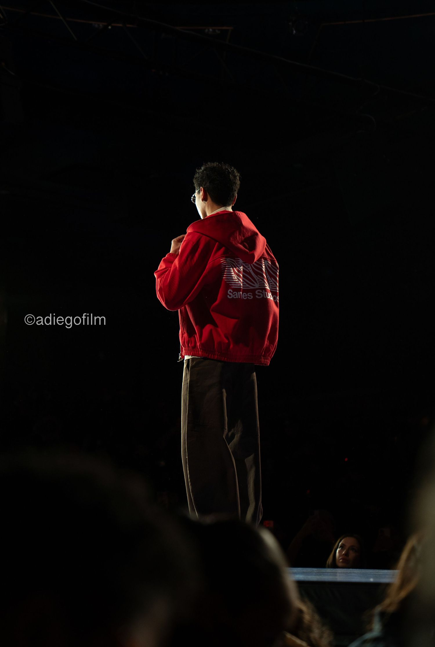 Performer in red jacket stands on dark stage with dramatic backlighting.