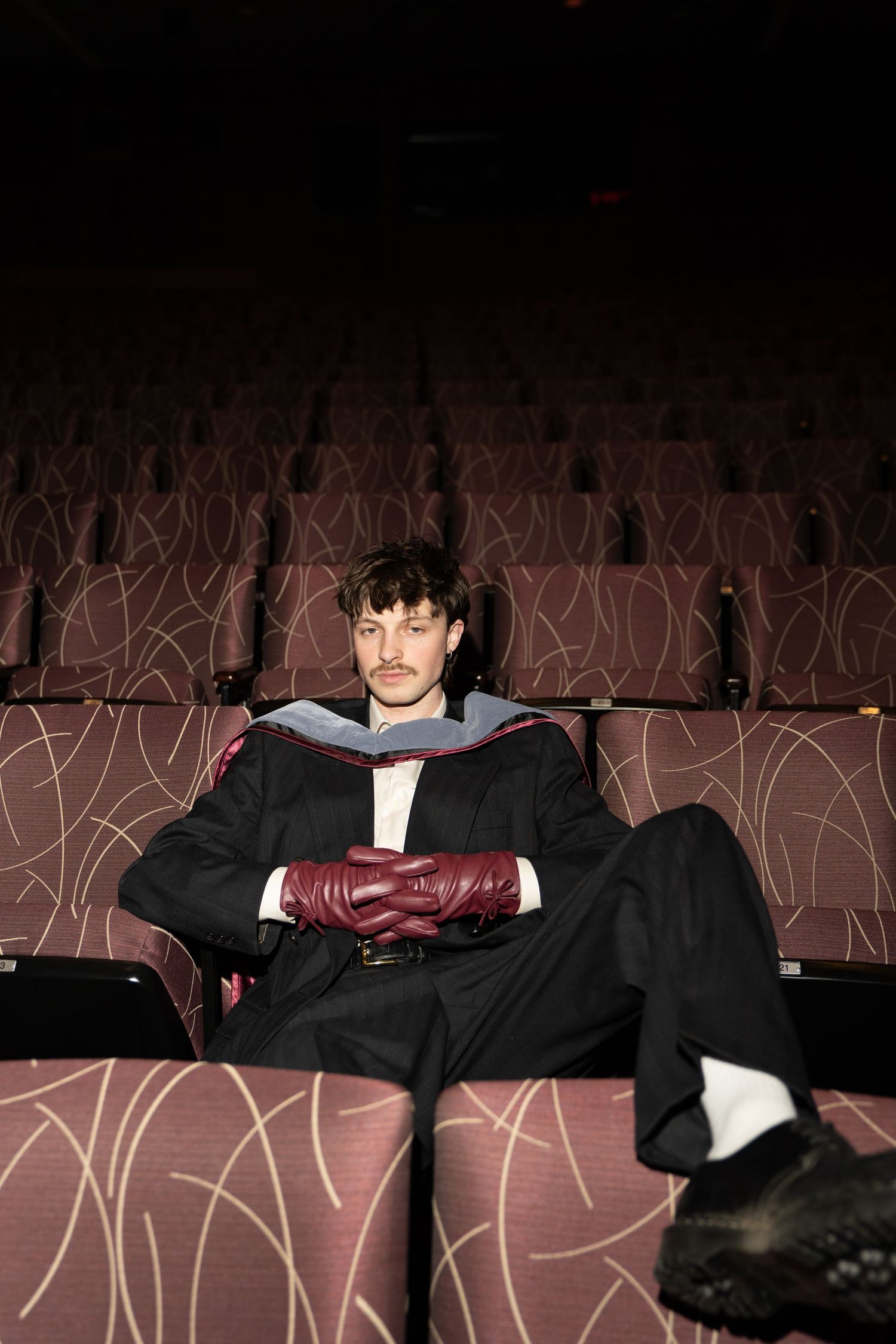 Person in formal attire reclining across multiple theater seats in a dark auditorium with patterned walls.