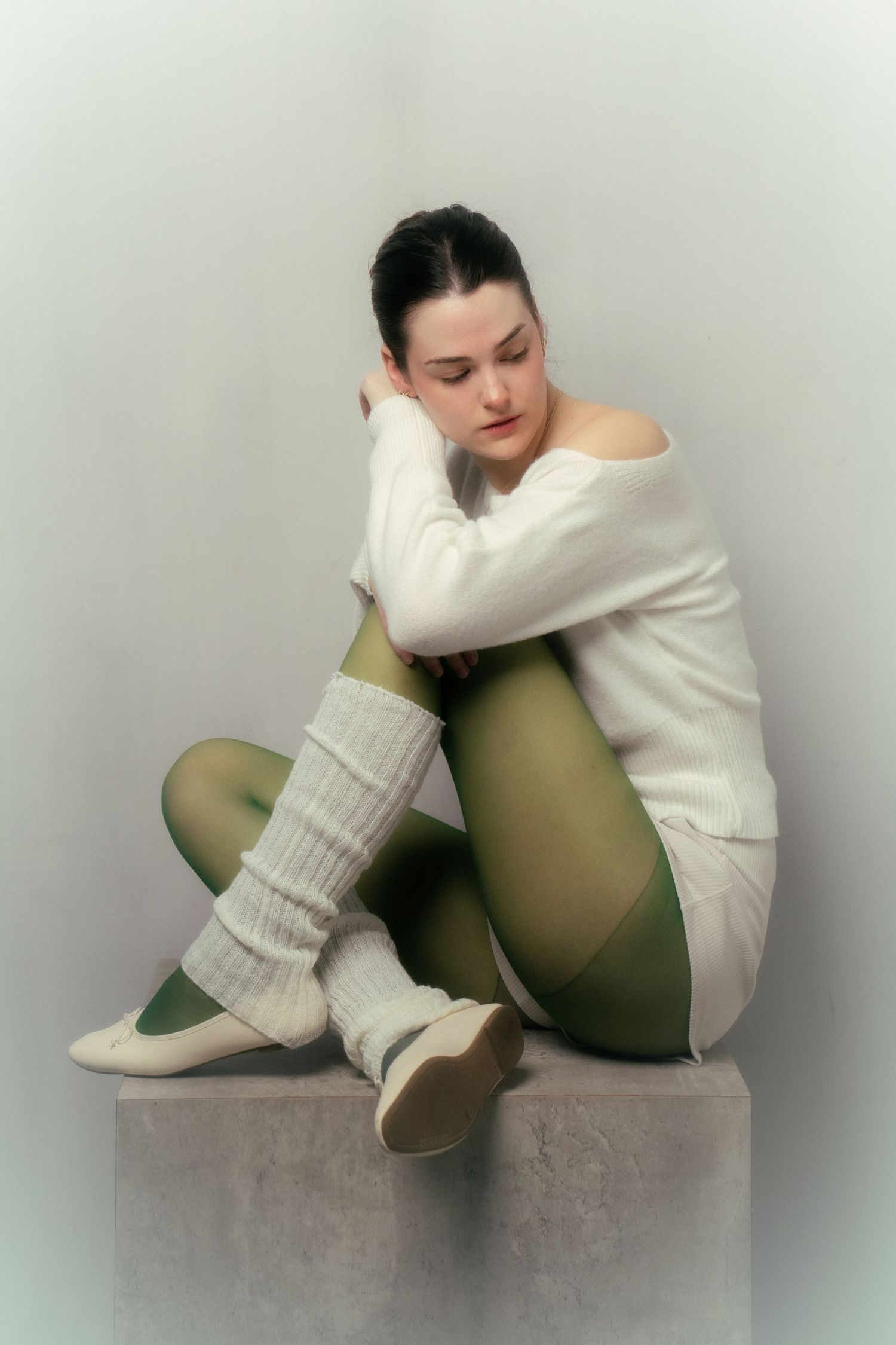 Figure in white sweater sits curled up against olive green cushion in minimalist artistic portrait series.