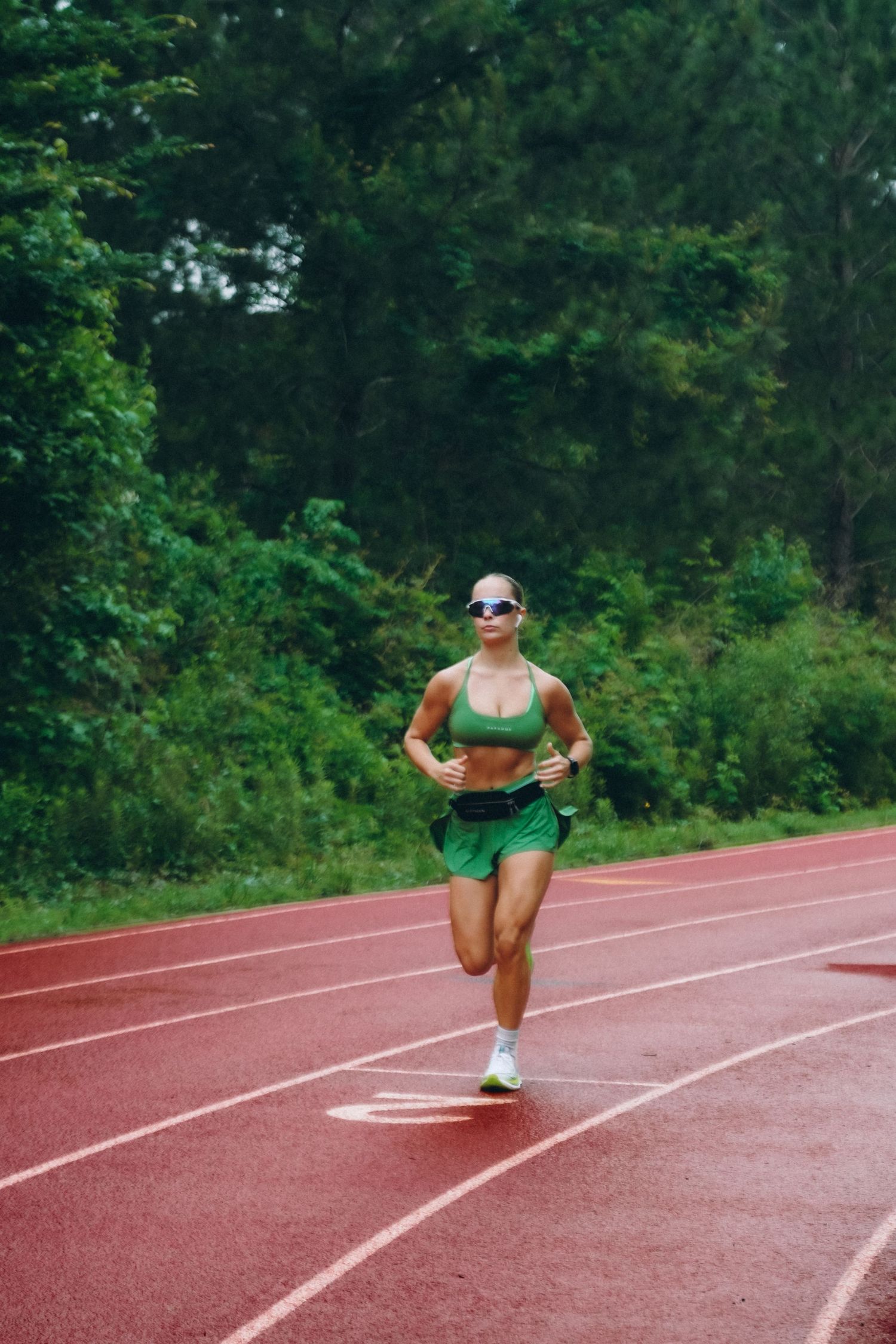 A fit woman in green athletic wear runs on a red track surrounded by lush green trees.
