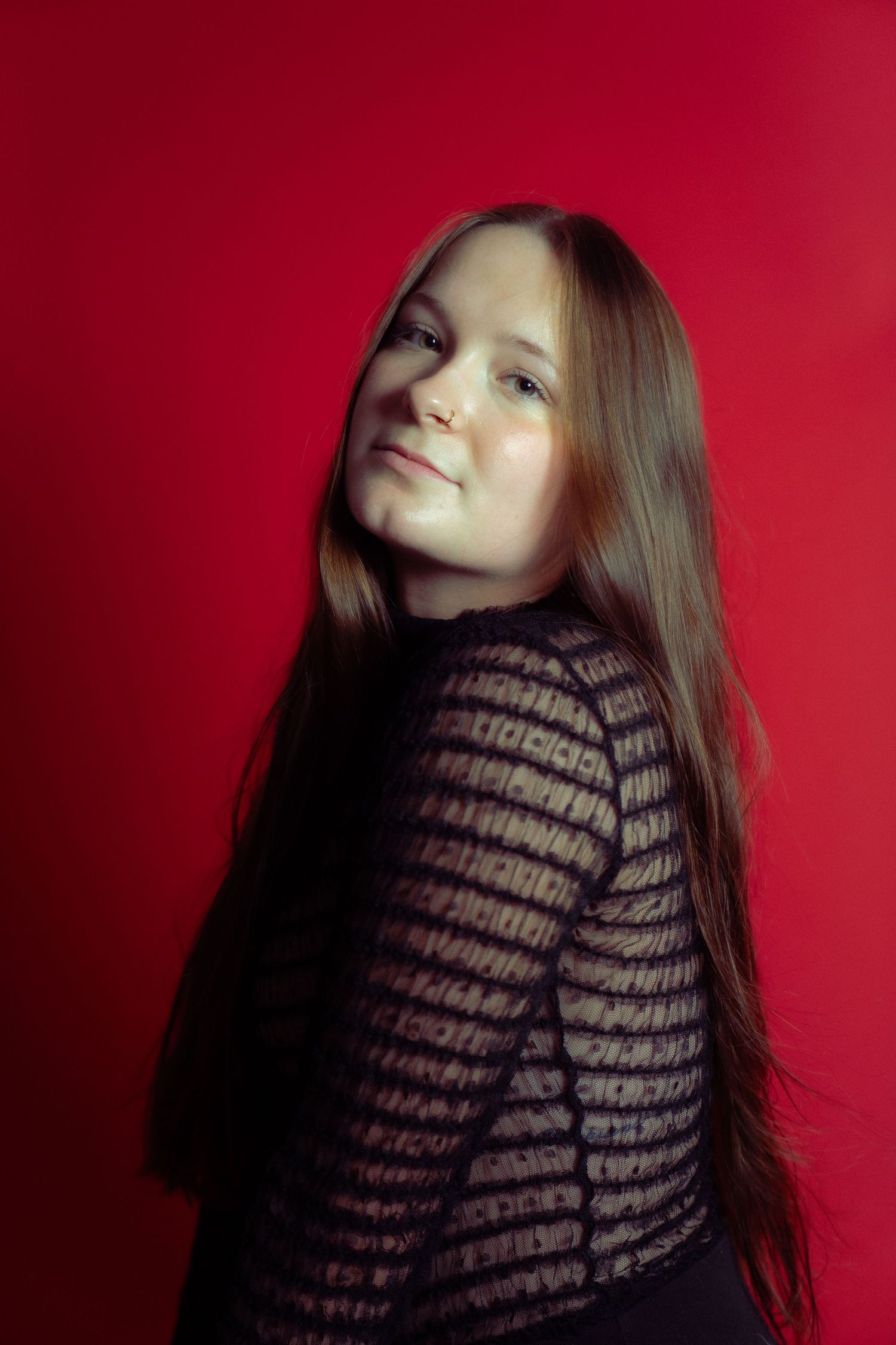 Portrait against red background showing long straight brown hair and sheer striped black top in moody lighting.
