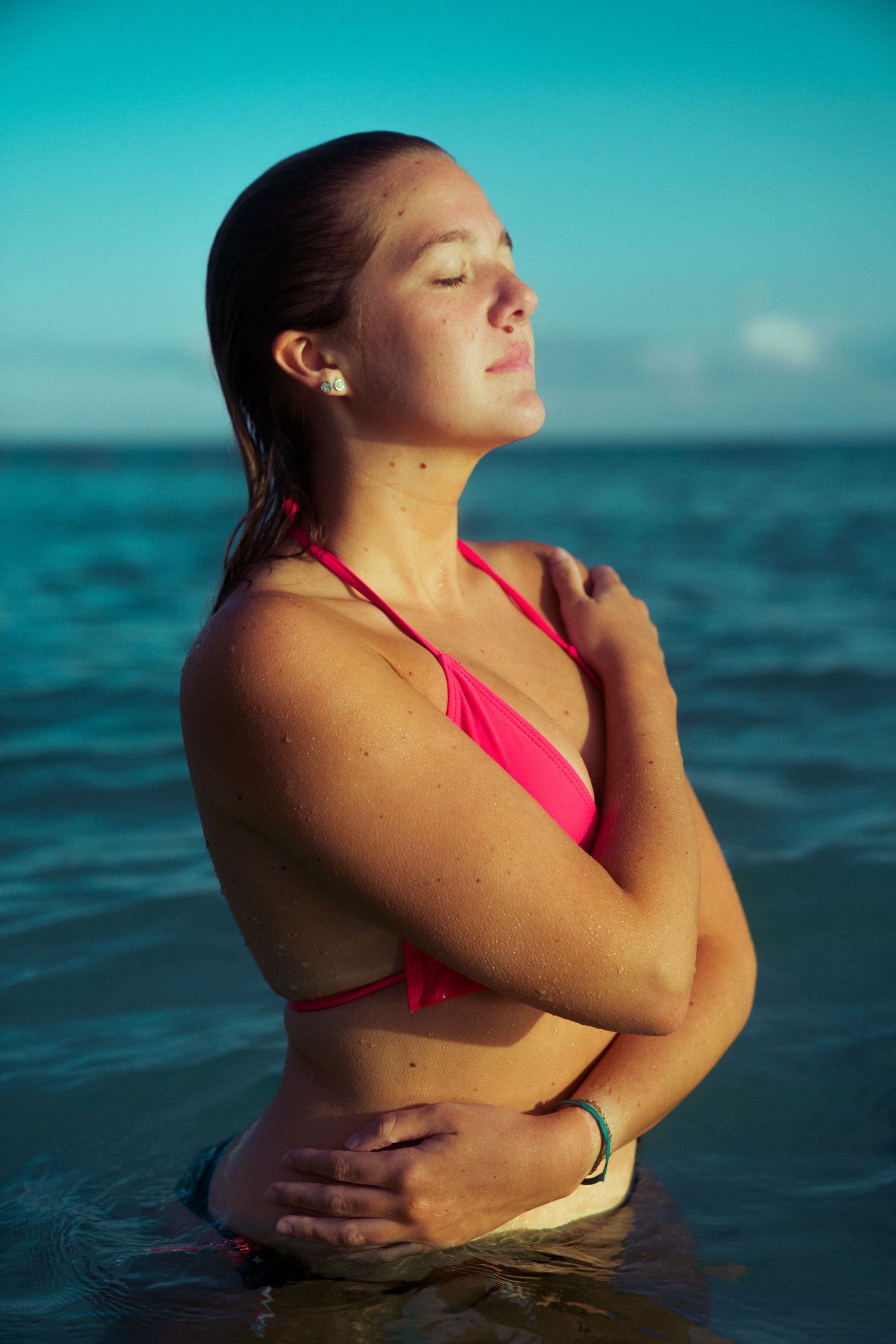 A person in a pink swimsuit stands peacefully in turquoise ocean waters during sunset with eyes closed.