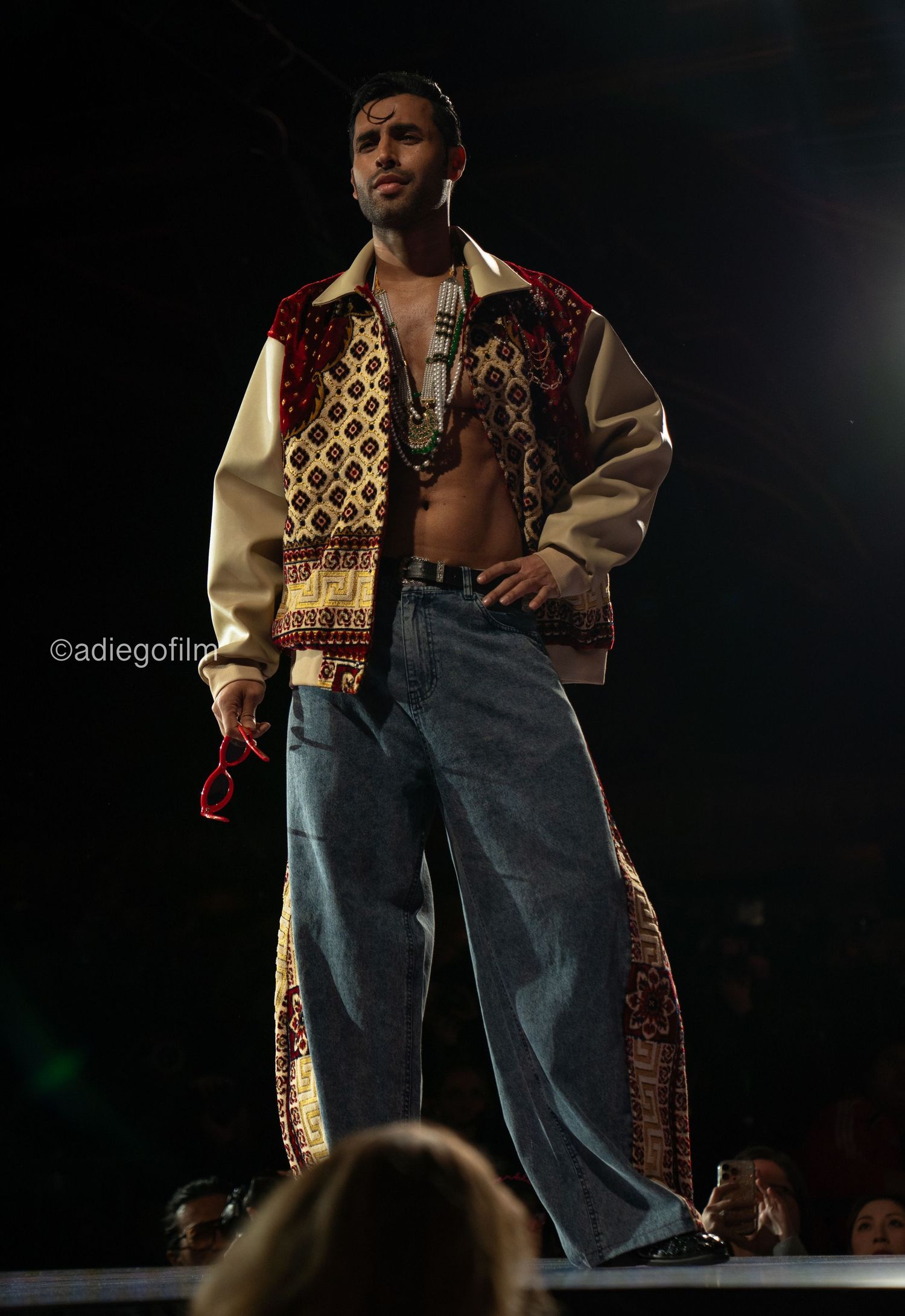 Performer wearing baggy jeans and patterned vest strikes dramatic poses on dark stage during fashion show.