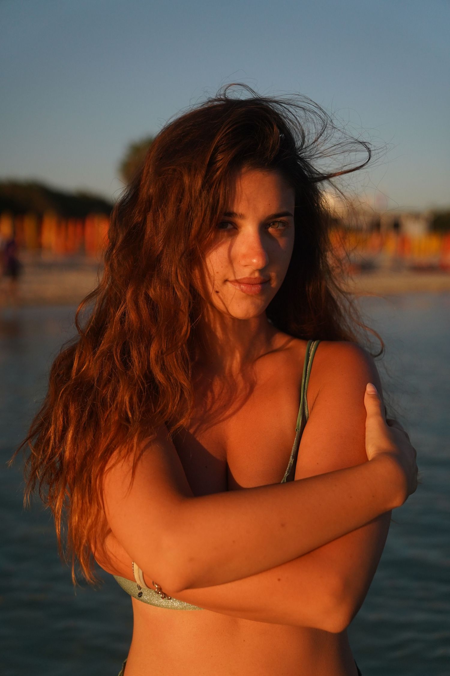 Person with long wavy red hair and tan skin poses at golden hour near water in swimwear.