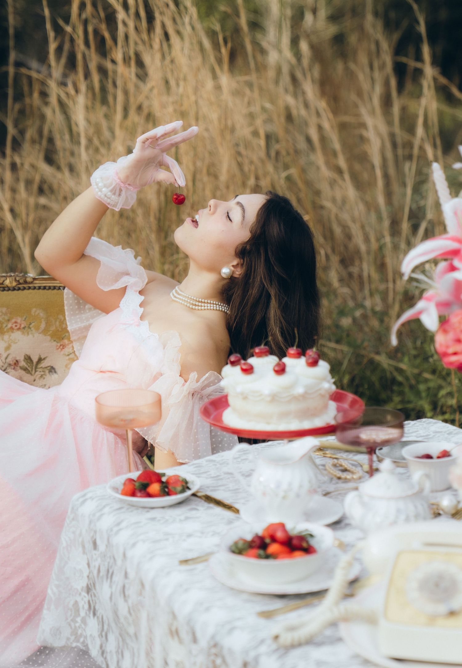 A whimsical tea party setup outdoors with pink dress, white tablecloth, strawberries and cake on a rustic table.