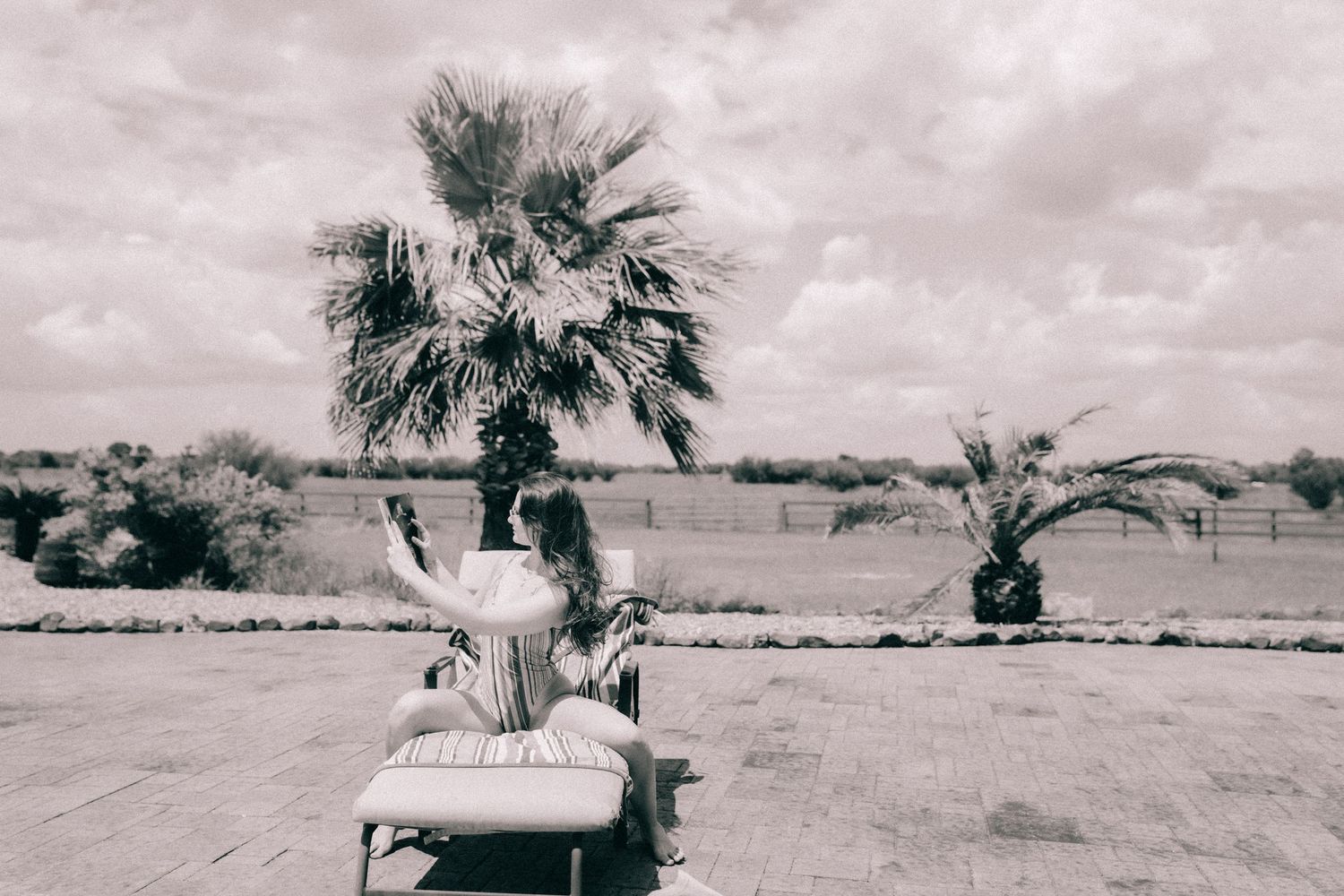 Person lounging on outdoor chair with palm tree in desert landscape, captured in monochrome style.