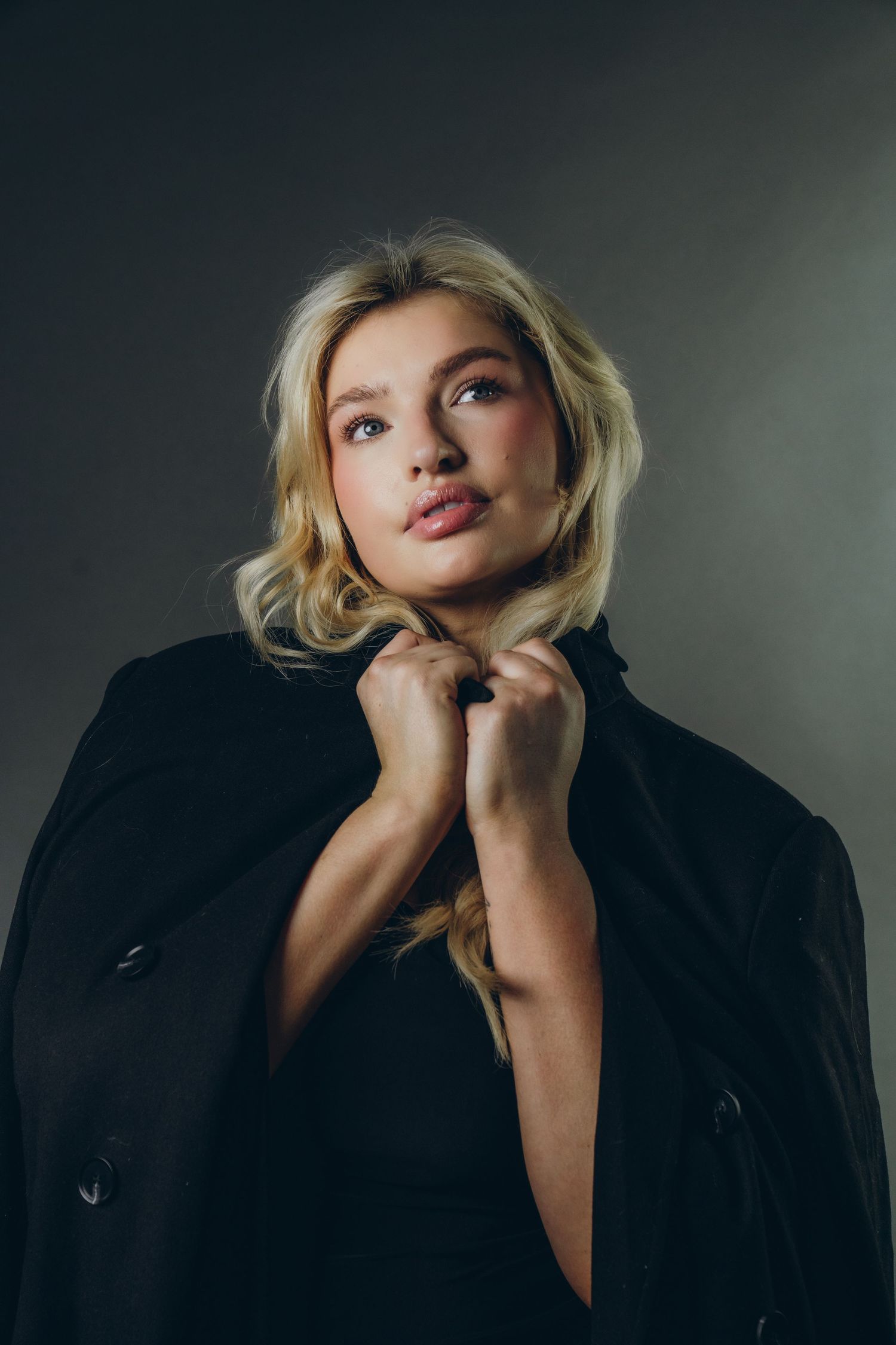 Blonde woman in black coat holding collar, studio portrait with dark background.