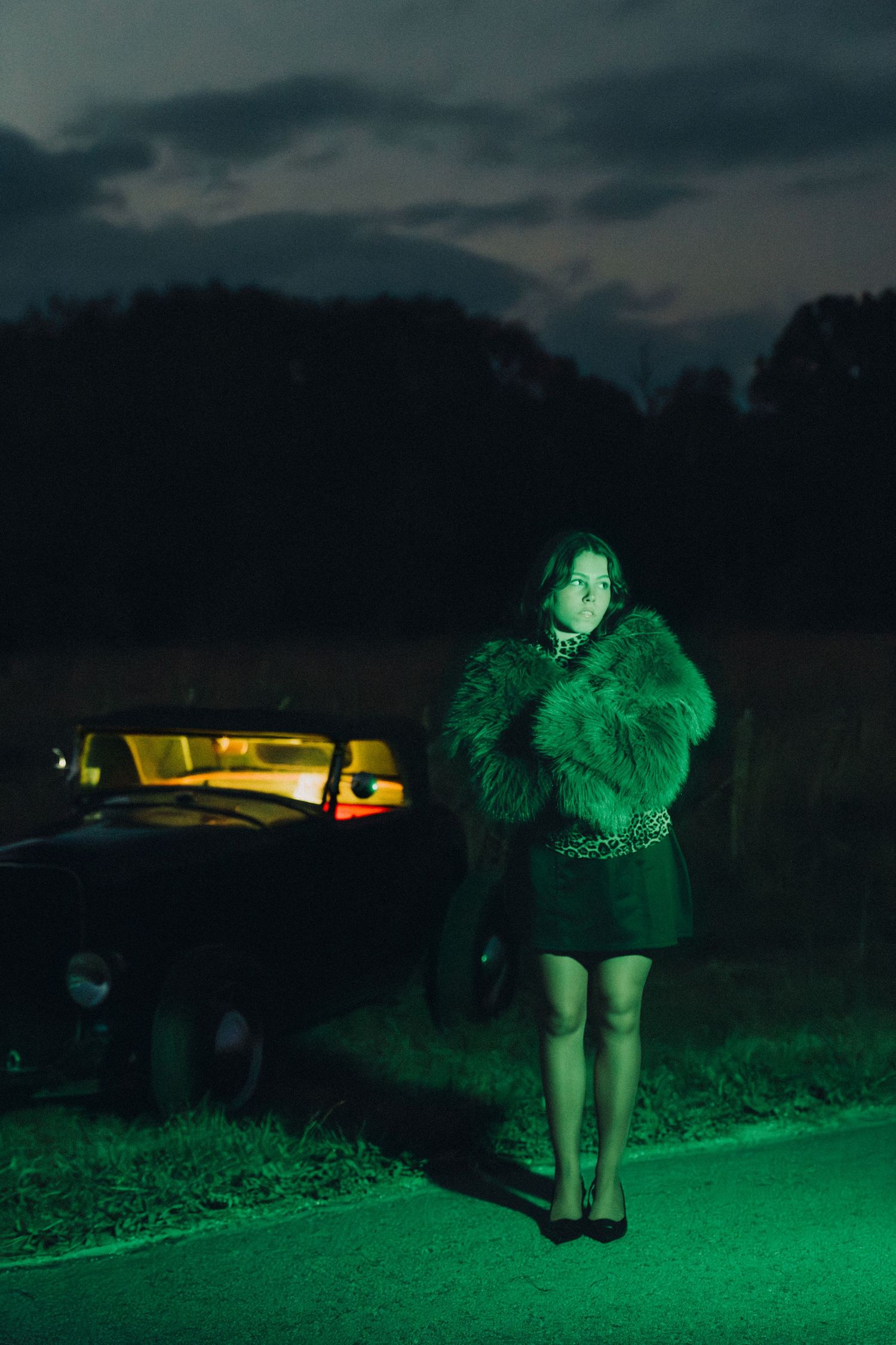 A figure in a fur coat stands illuminated by eerie green lighting next to a car at night on a dark road.