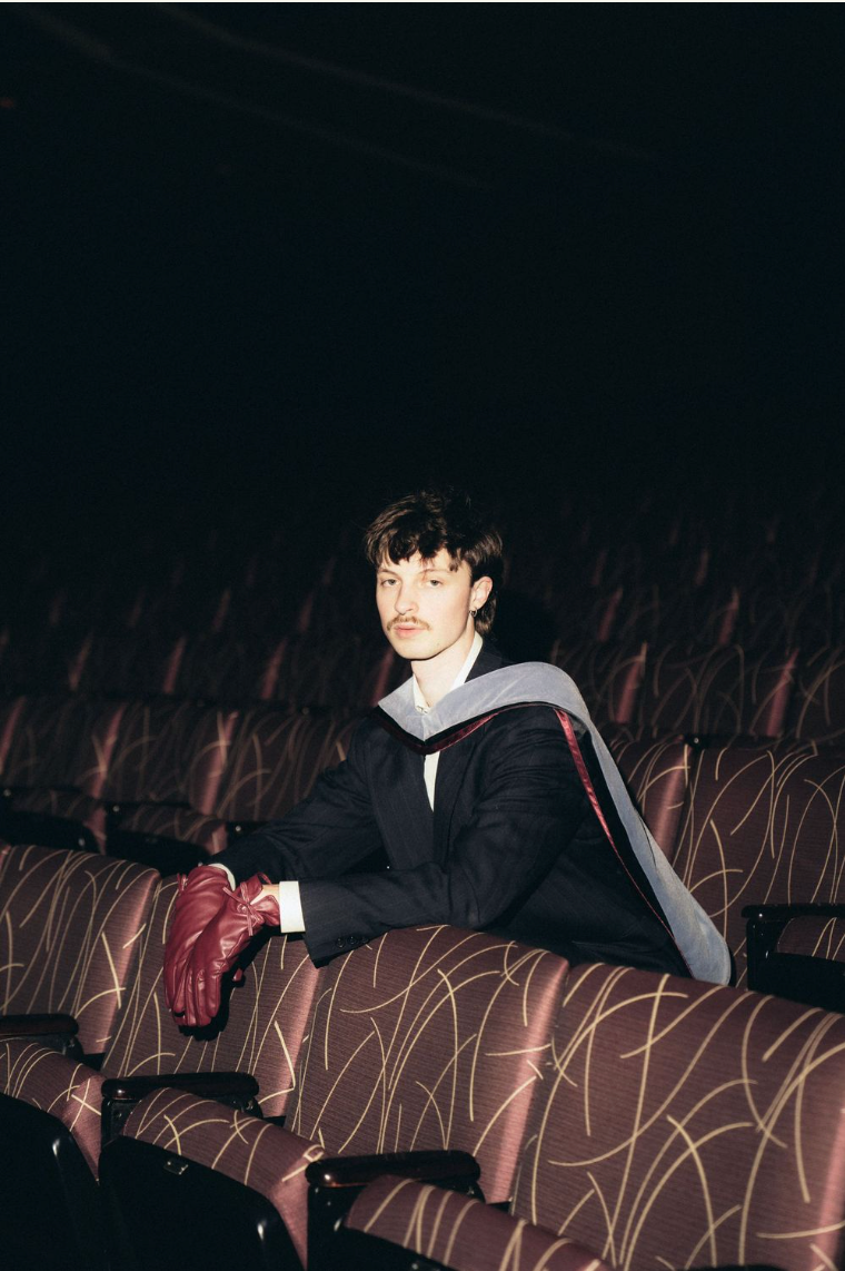 A person sits in an empty theater auditorium with rows of unoccupied seats visible in the dark lighting.