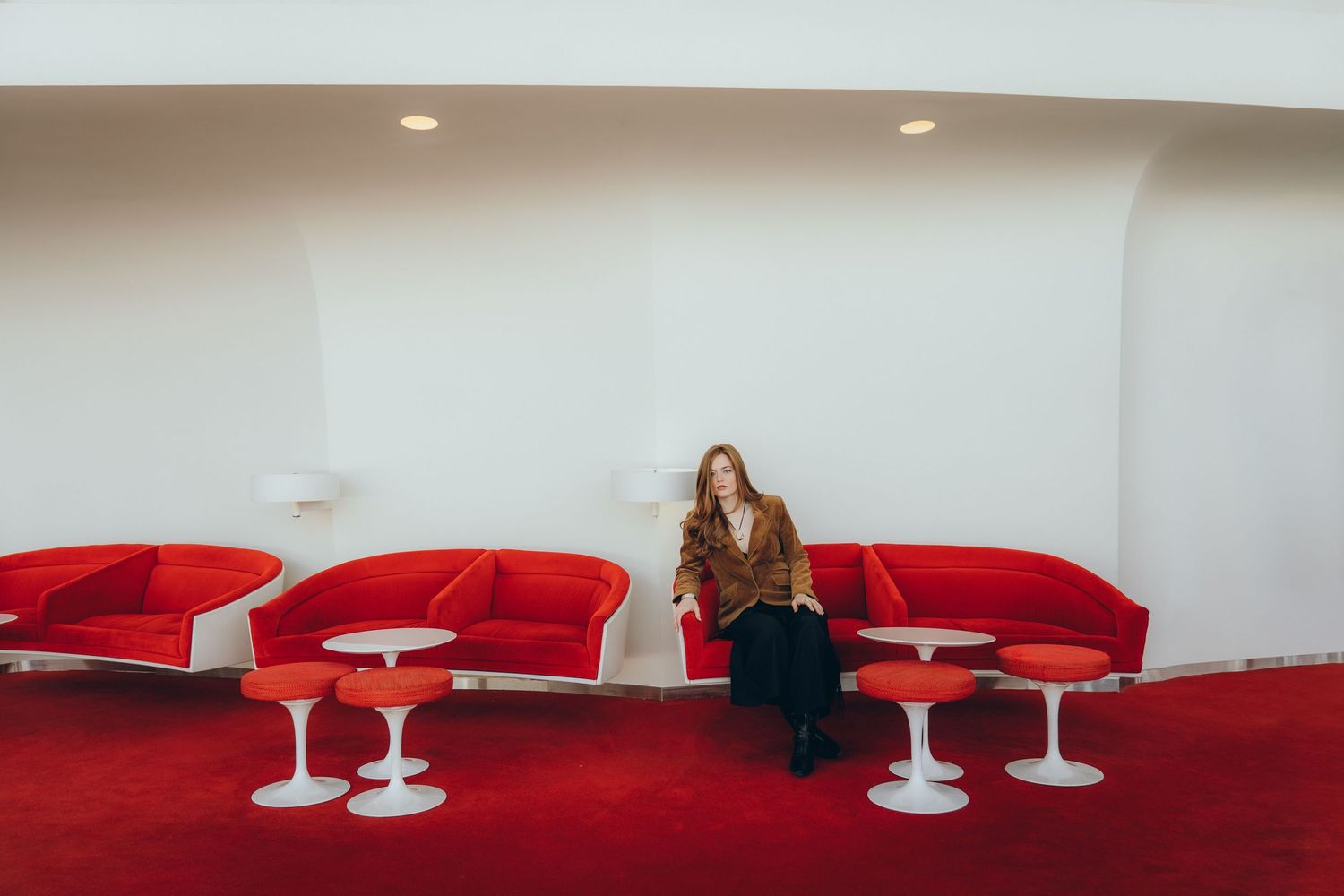 Someone sits alone in a retro-modern waiting area with bright red chairs and white tulip-style bases on red carpet.