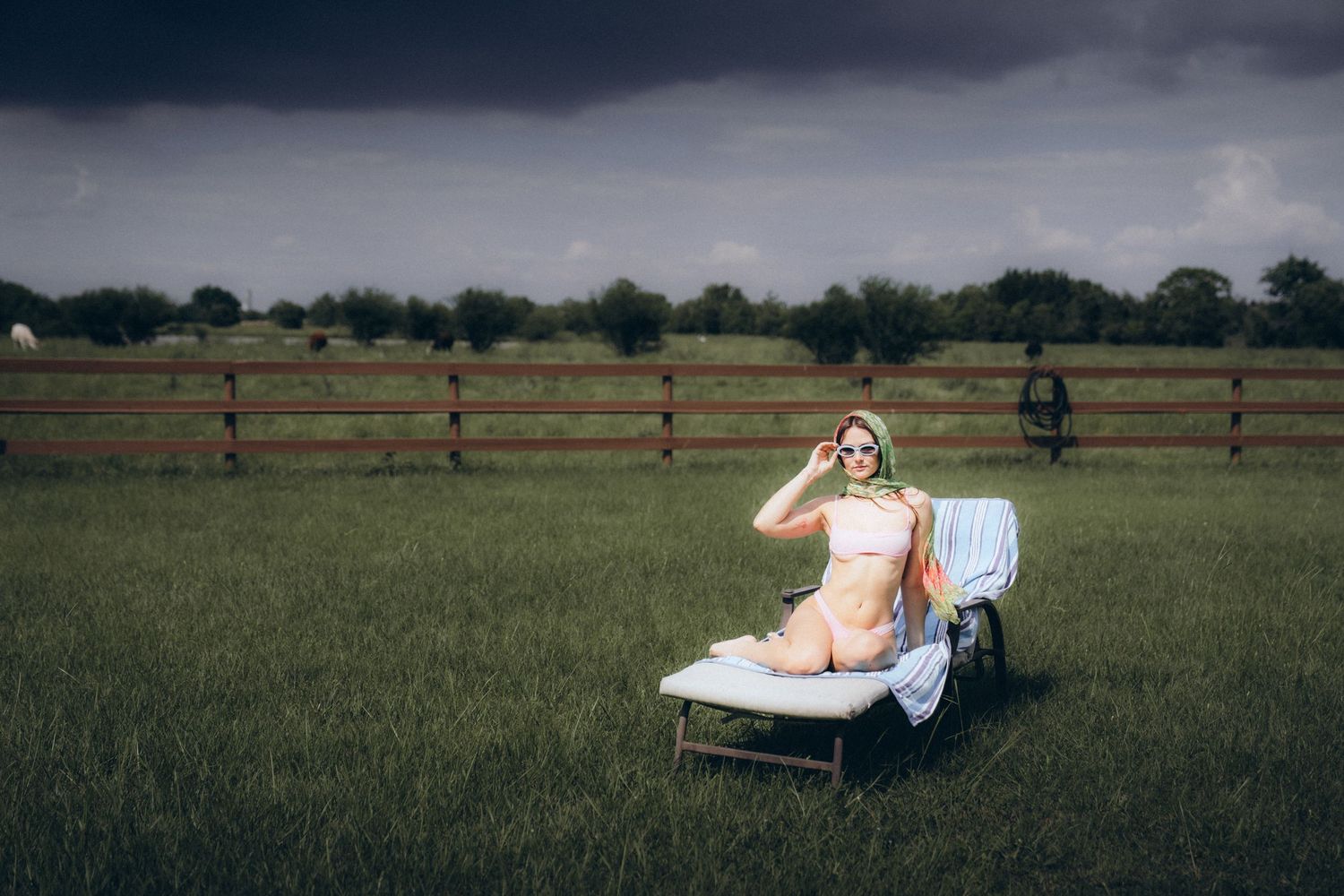A person relaxes on a white lounge chair in a grassy field with wooden fences in the background.