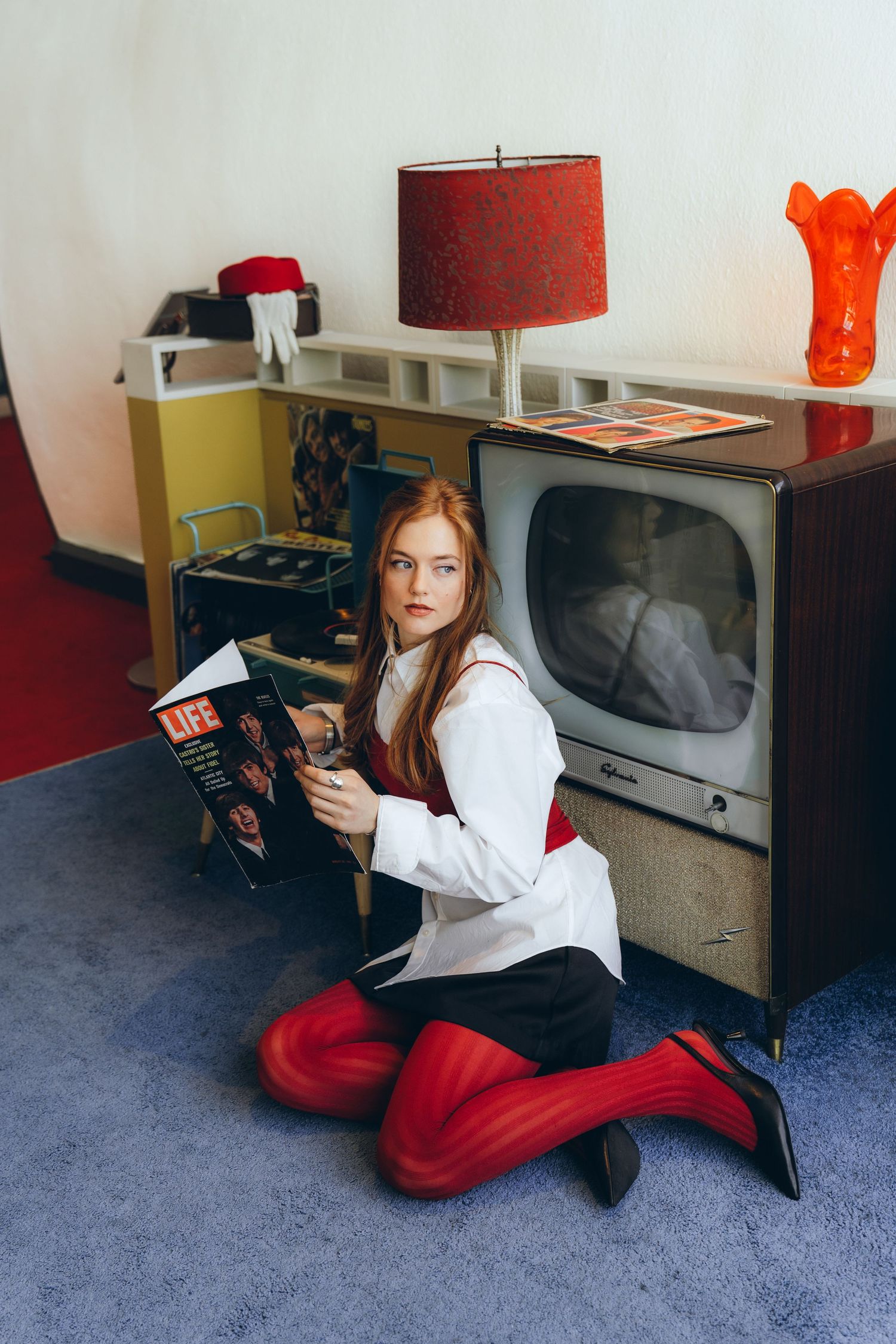 A person in a white shirt and red pants lies on blue carpet while holding a camera in a room with orange decor.
