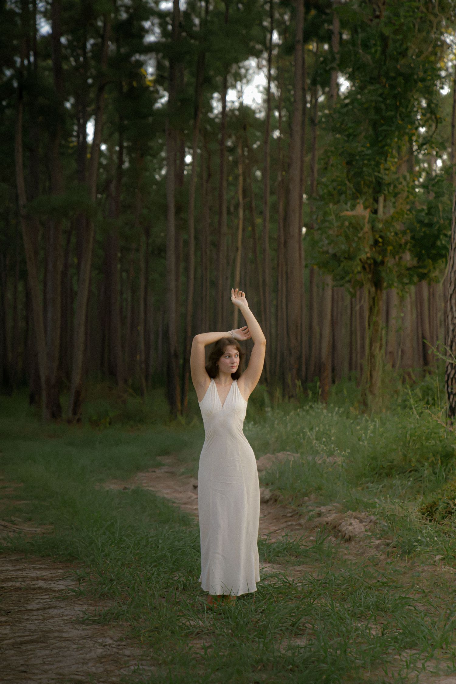 Figure in white dress raises arms gracefully on forest path surrounded by tall pine trees.