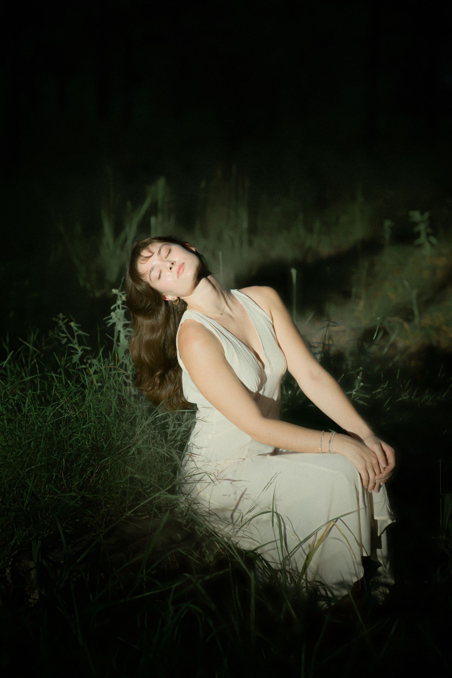 Dramatic nighttime portrait of figure in white dress posed among tall grasses.
